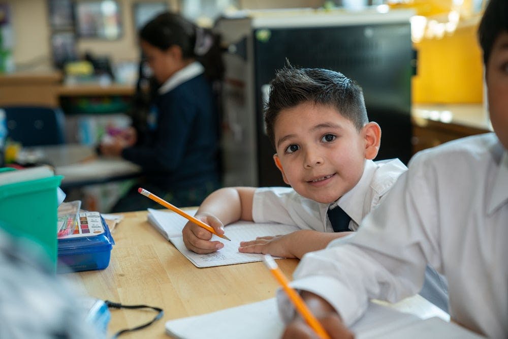 Child writing in a notebook at a desk in a classroom, looking at the camera