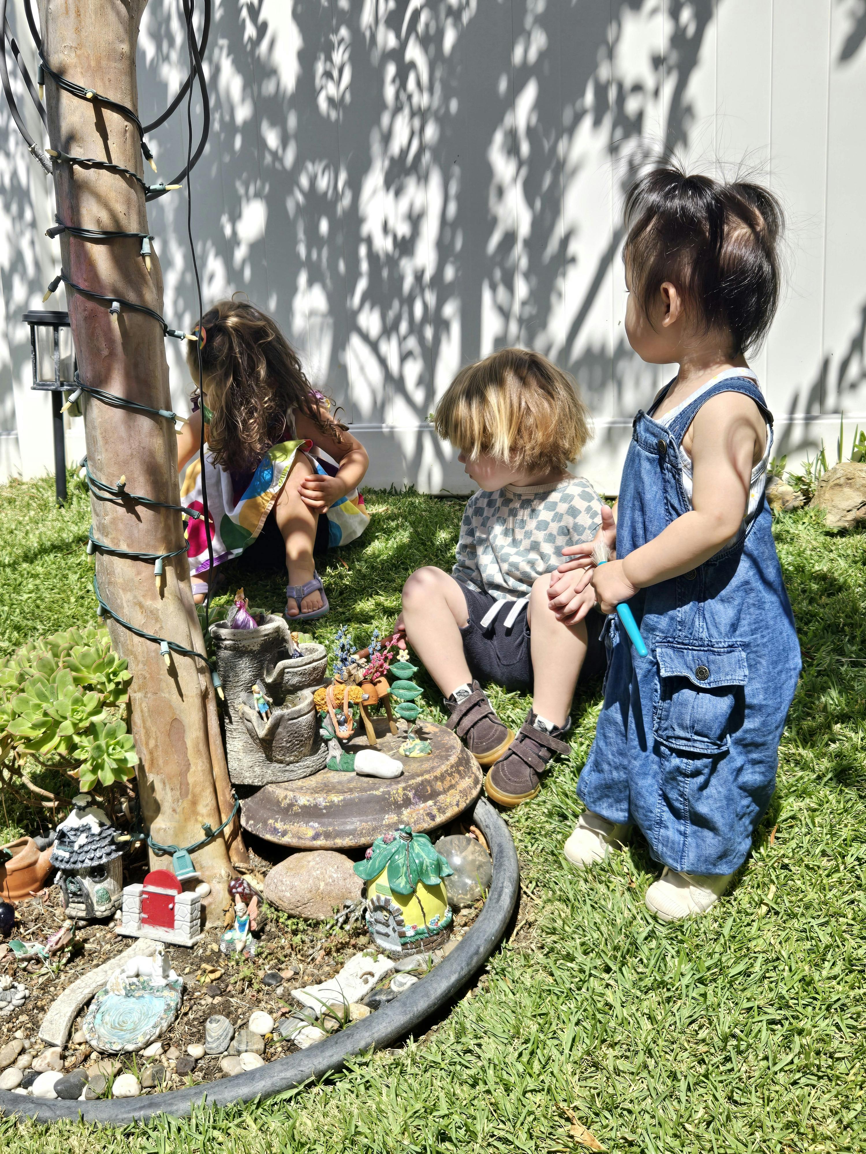 Three children playing with a fairy garden around a tree trunk