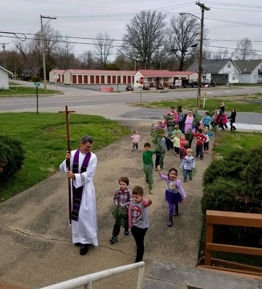 Person in white robe holding a cross leading a group of children