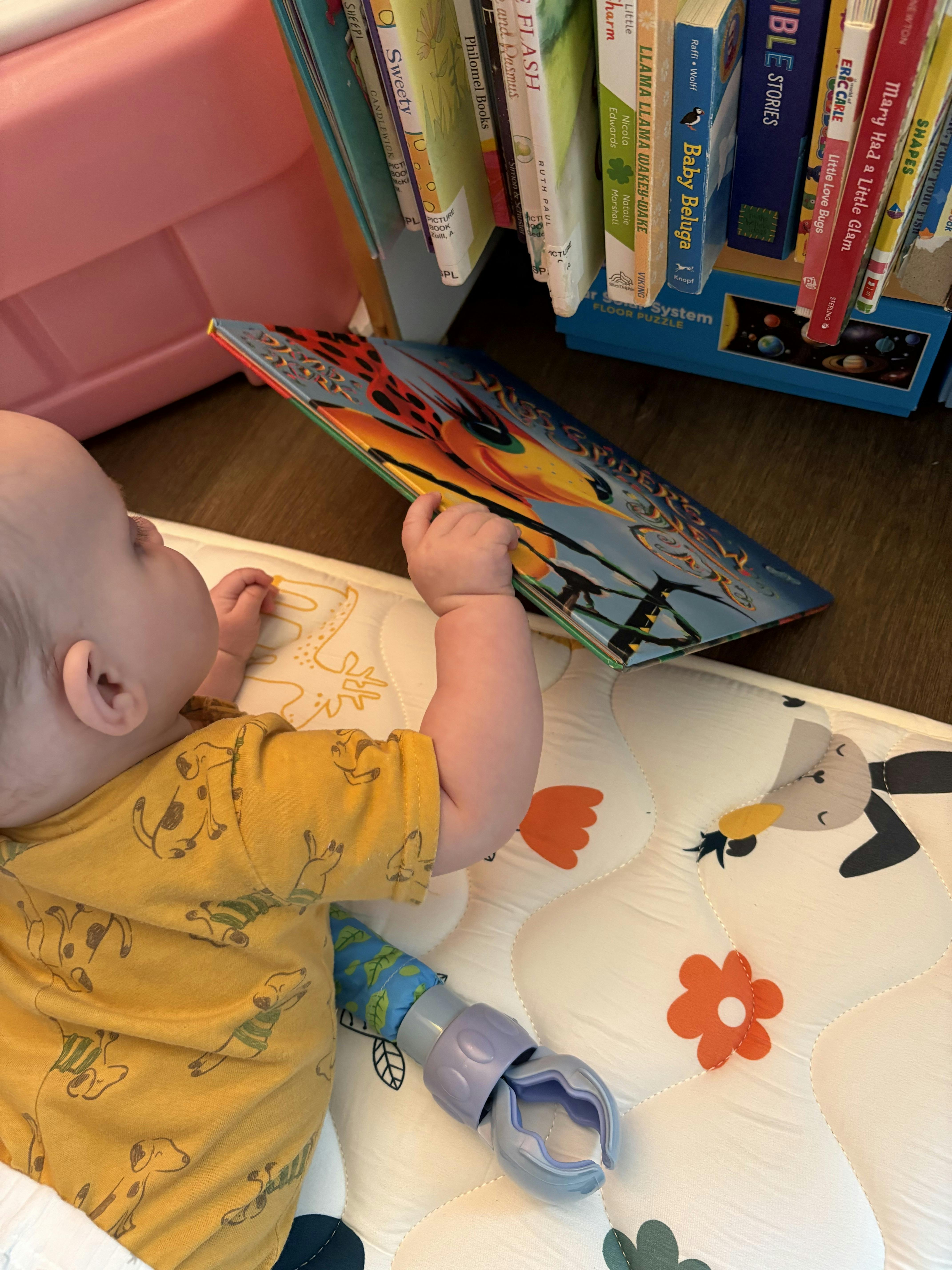 Baby lying on a playmat, reaching for a colorful book titled "Miss Spider"