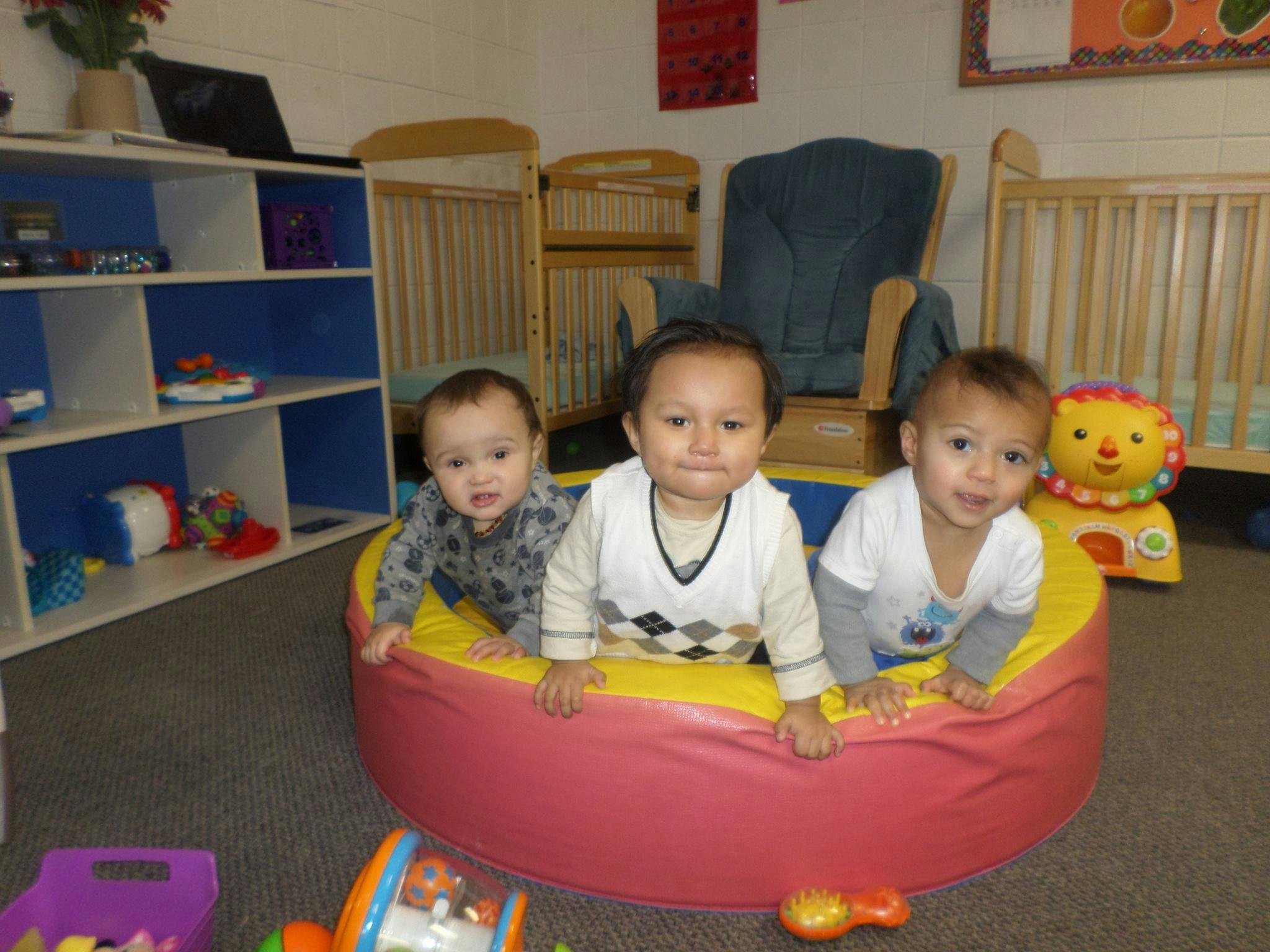 Three babies in a soft play pit in a room with cribs