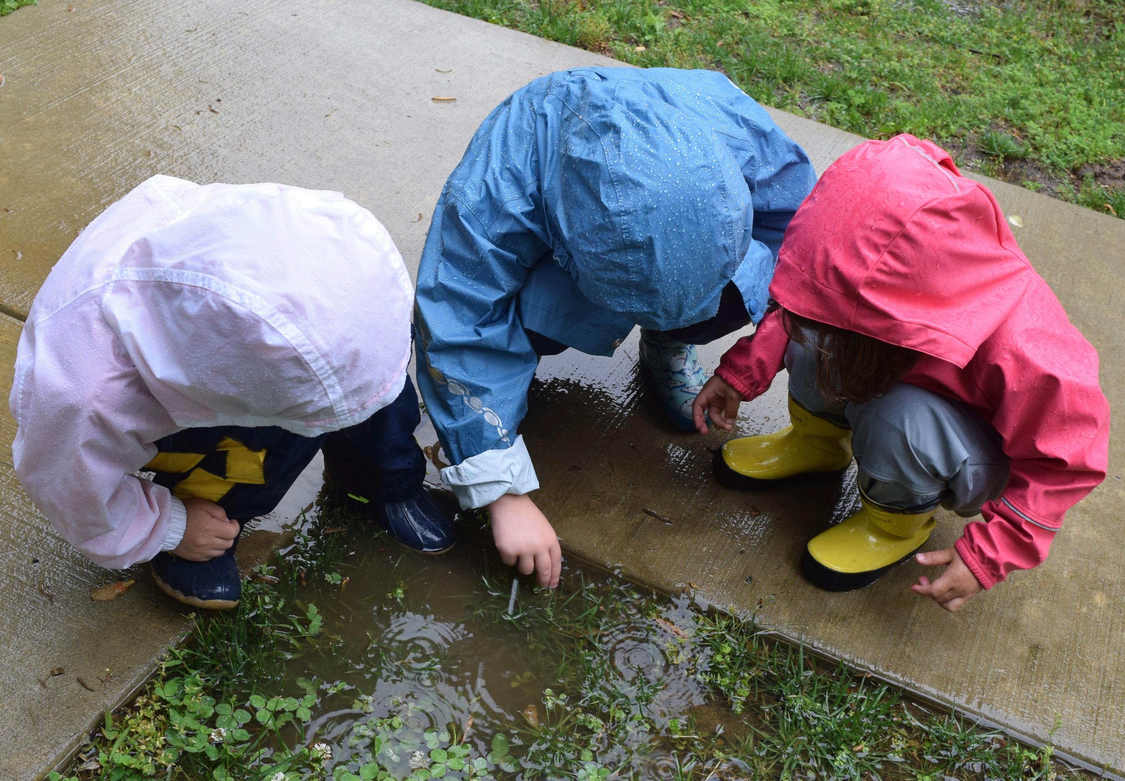 Three children in rain gear crouching around a puddle