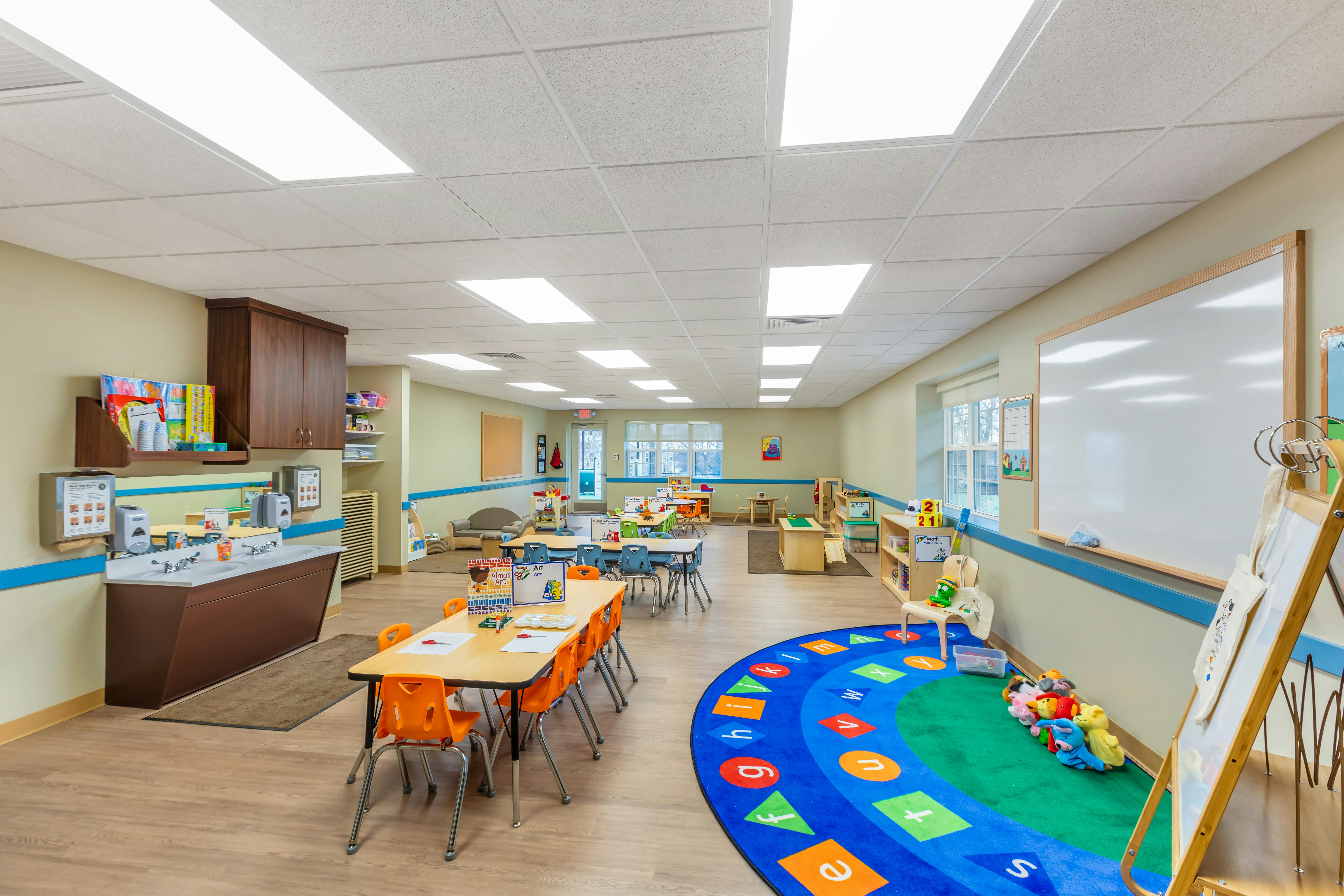 Classroom with child-sized tables and chairs, a sink station, and a colorful alphabet rug