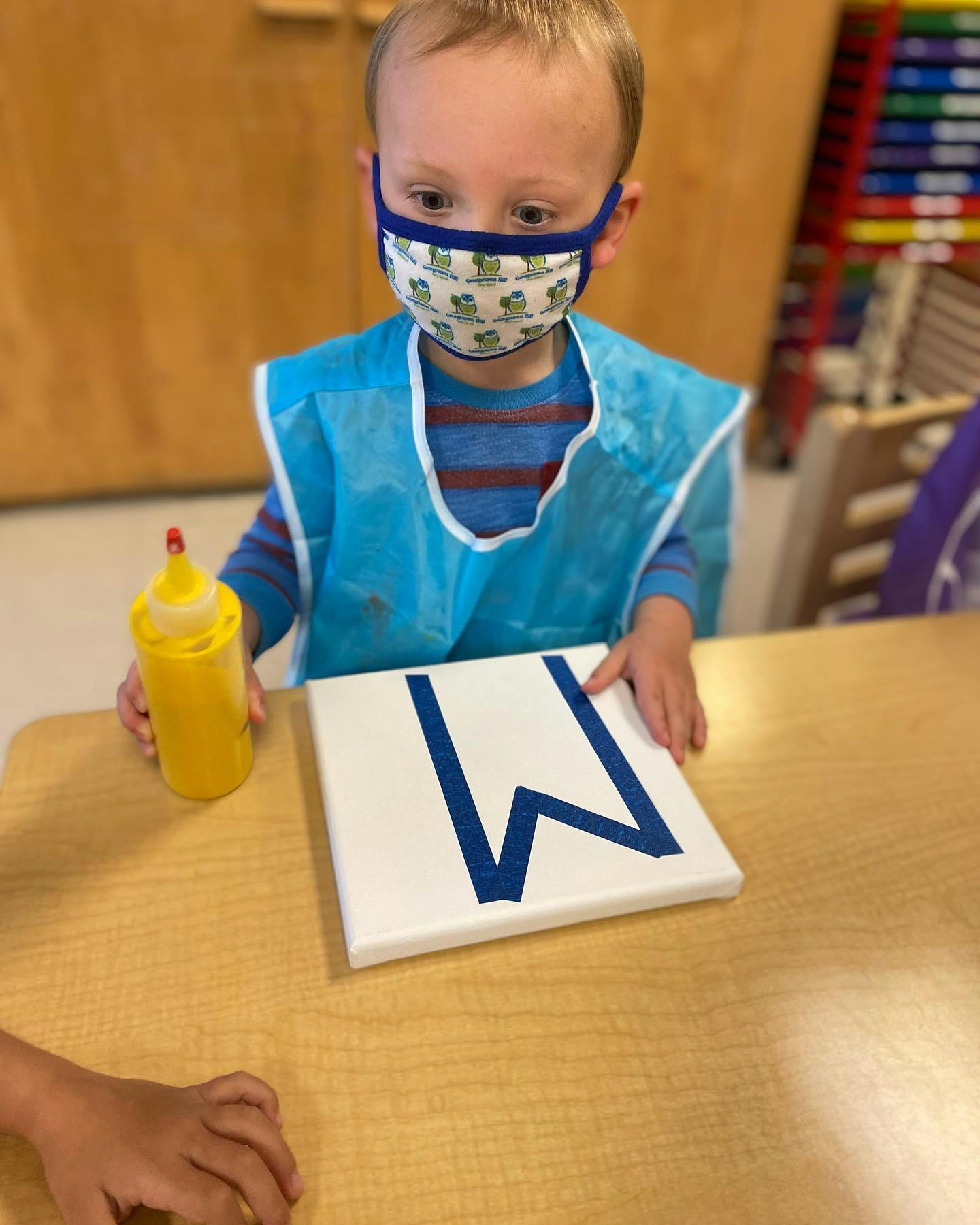 Child in a mask and smock holding a paint bottle and a canvas with a W