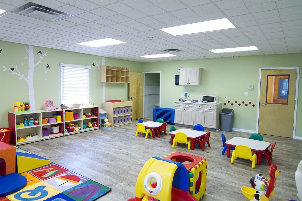 Empty classroom with small tables and chairs, shelves with toys, and a play mat