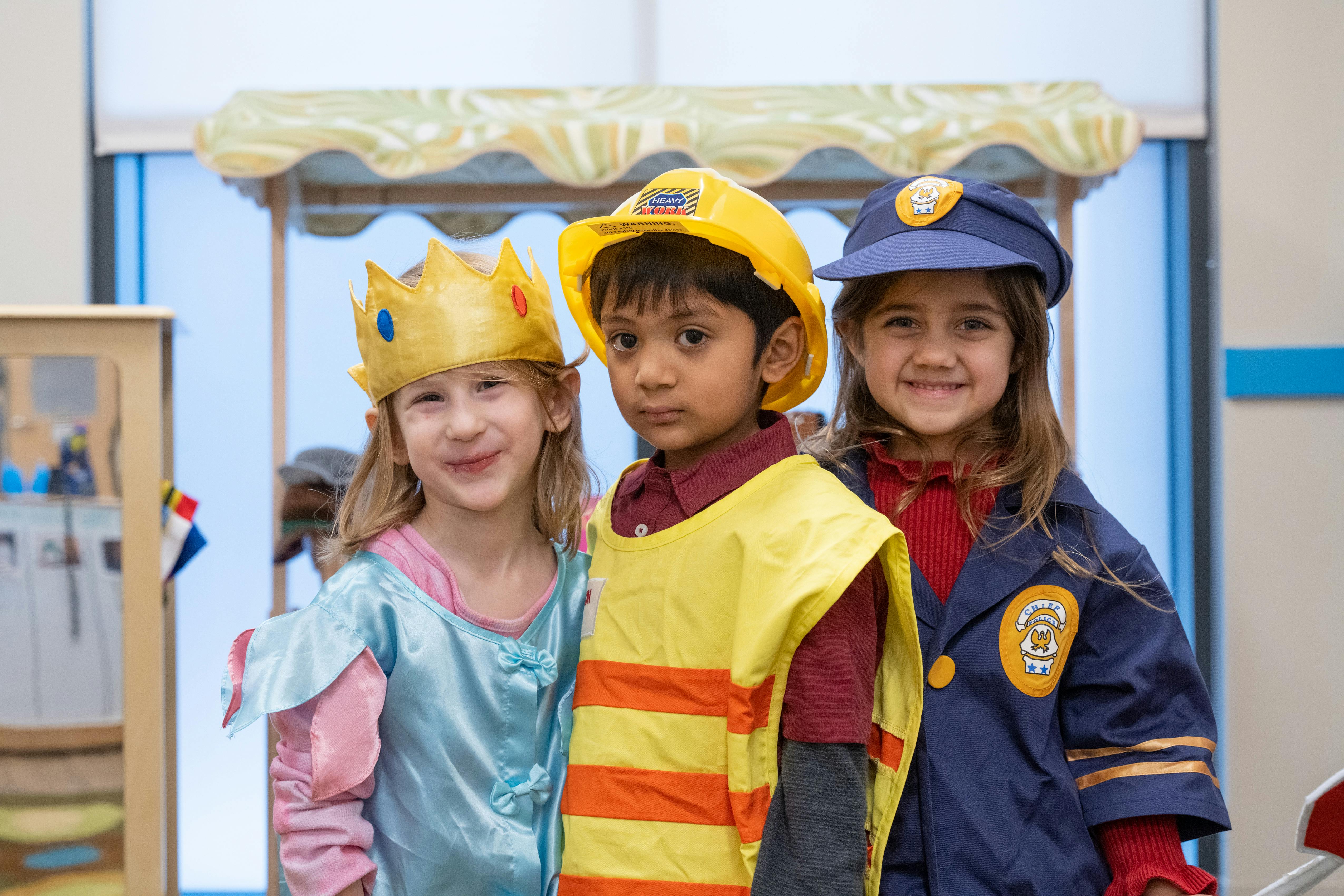 Three children in dress-up costumes, one in a crown, one in a construction hat, one in a police hat