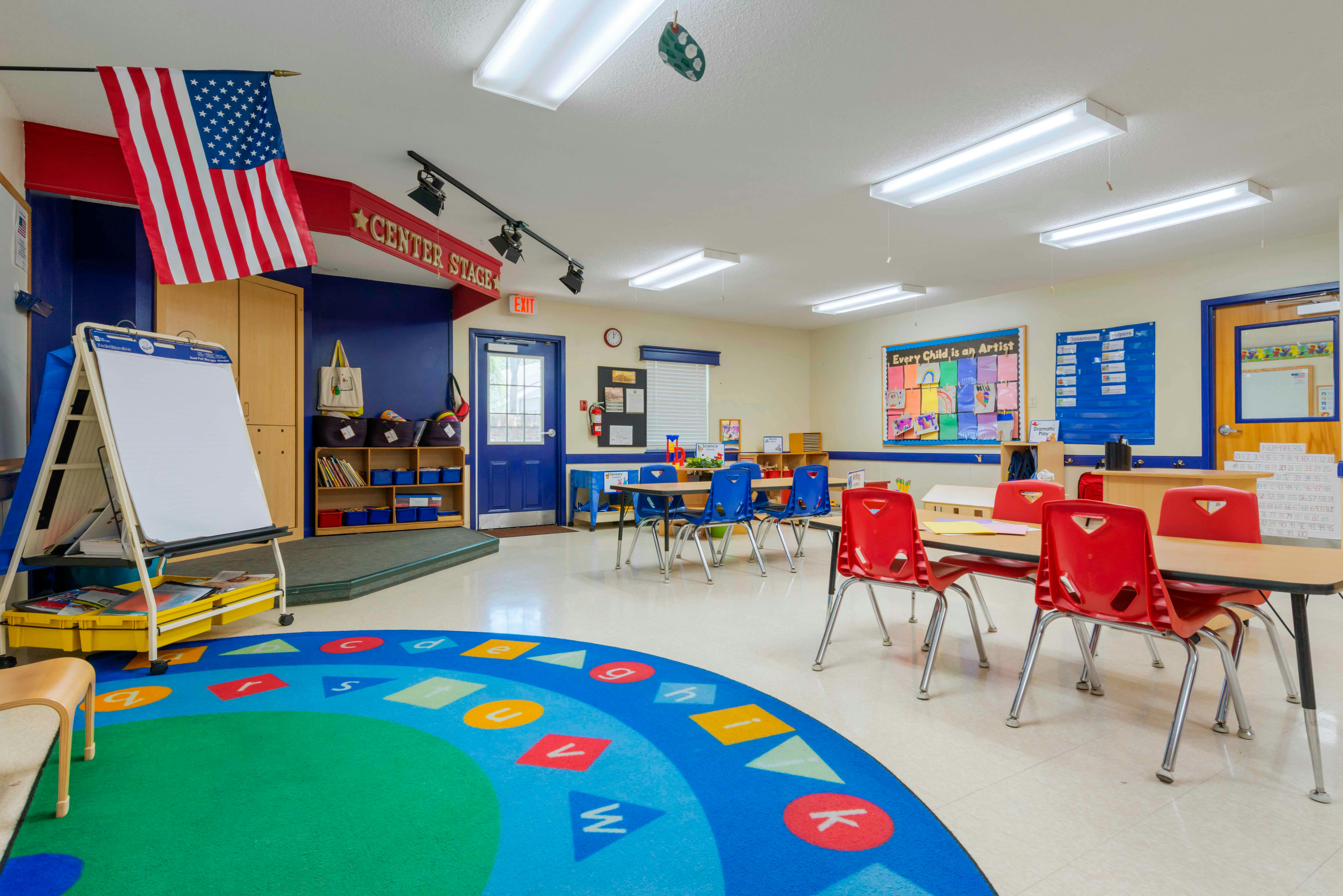 Empty classroom with small tables and chairs, a stage, and a circular rug with letters