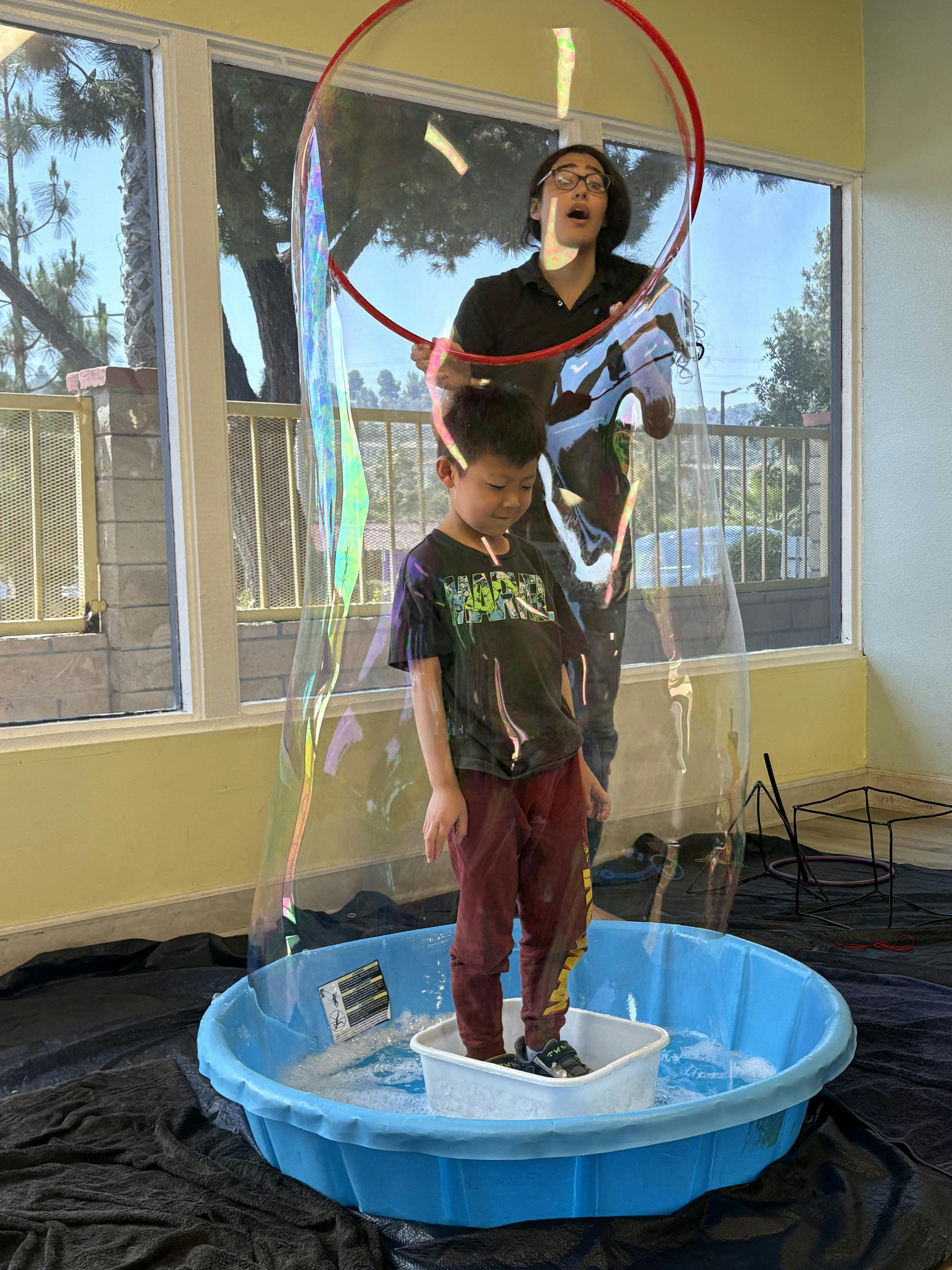 Child standing in a small pool, inside a giant bubble, with a person holding the bubble wand