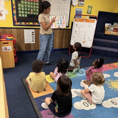 A person teaching numbers to children sitting on a classroom rug