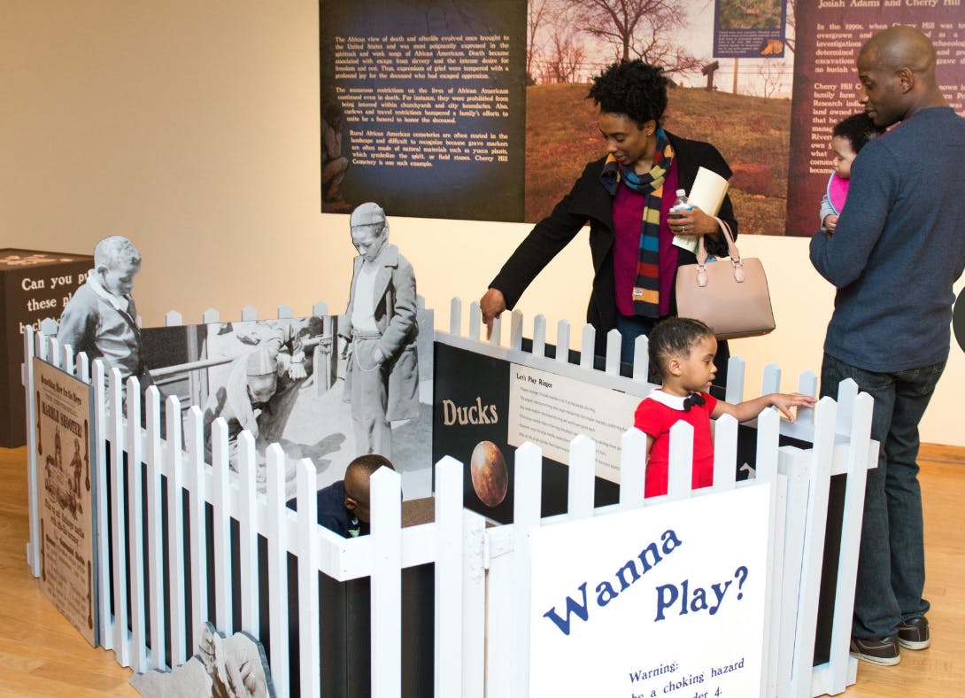 Adults and children interacting with an exhibit featuring a white picket fence