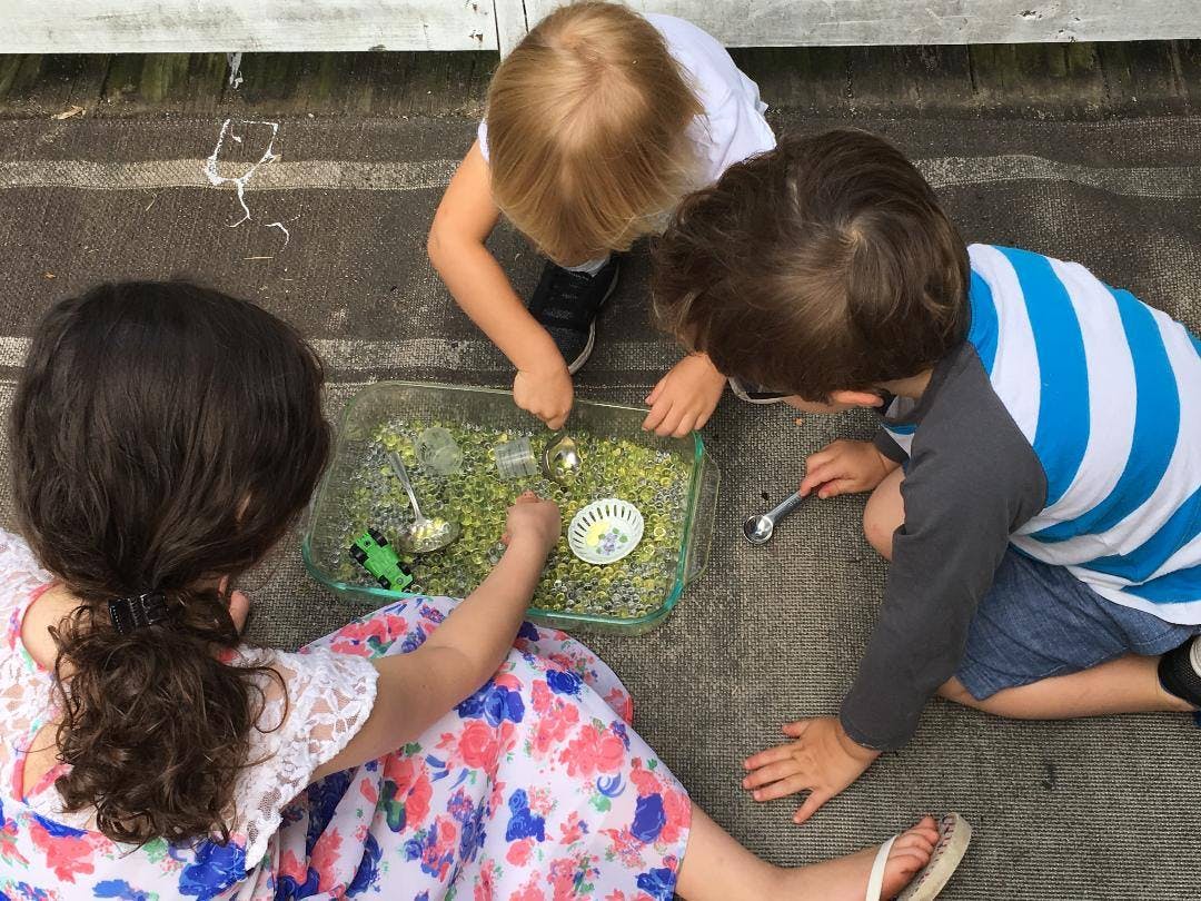 Three children playing with a sensory bin filled with green beads and spoons