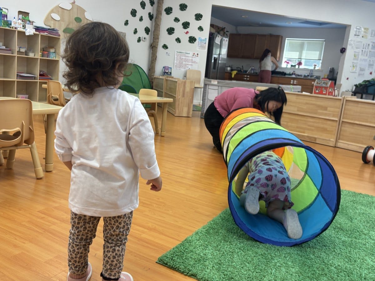 Child crawling through a play tunnel, adult watching in classroom