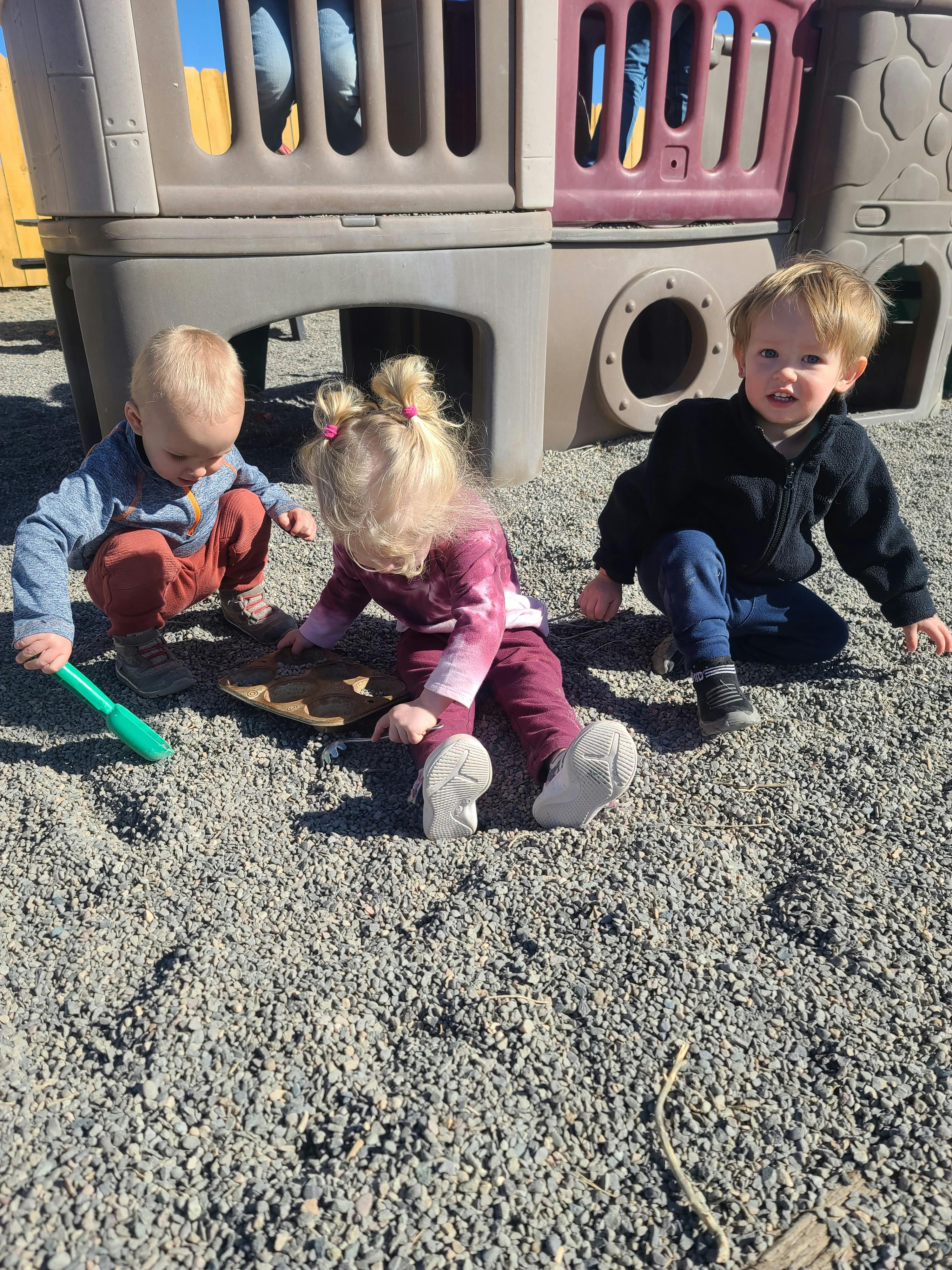 Three children playing in gravel with a toy shovel and pan
