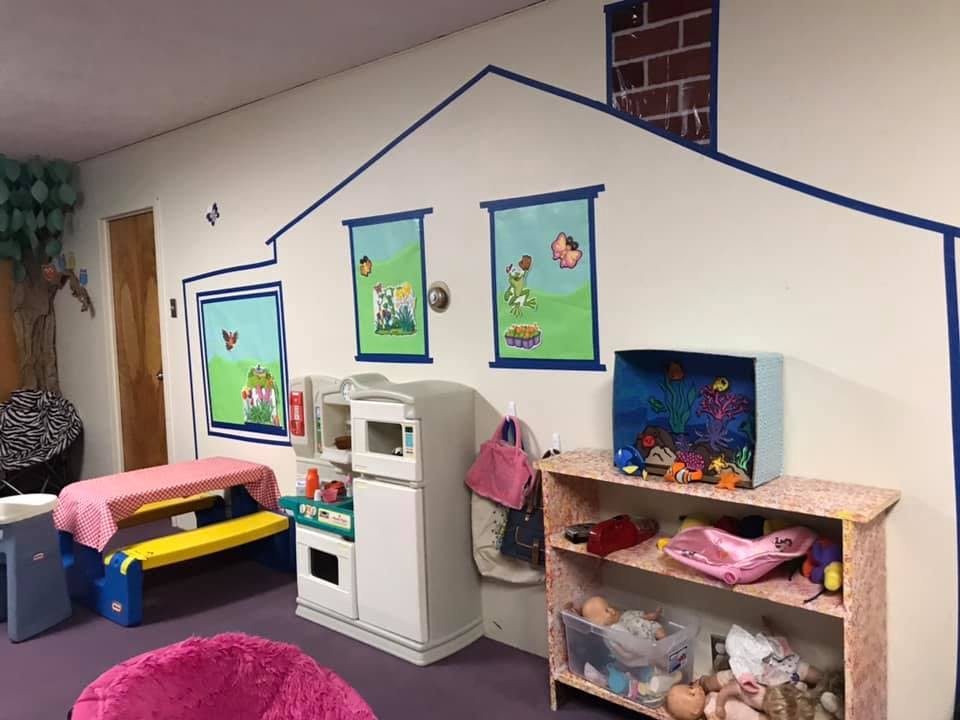 Playroom with toy kitchen, table, and shelves against a wall painted like a house