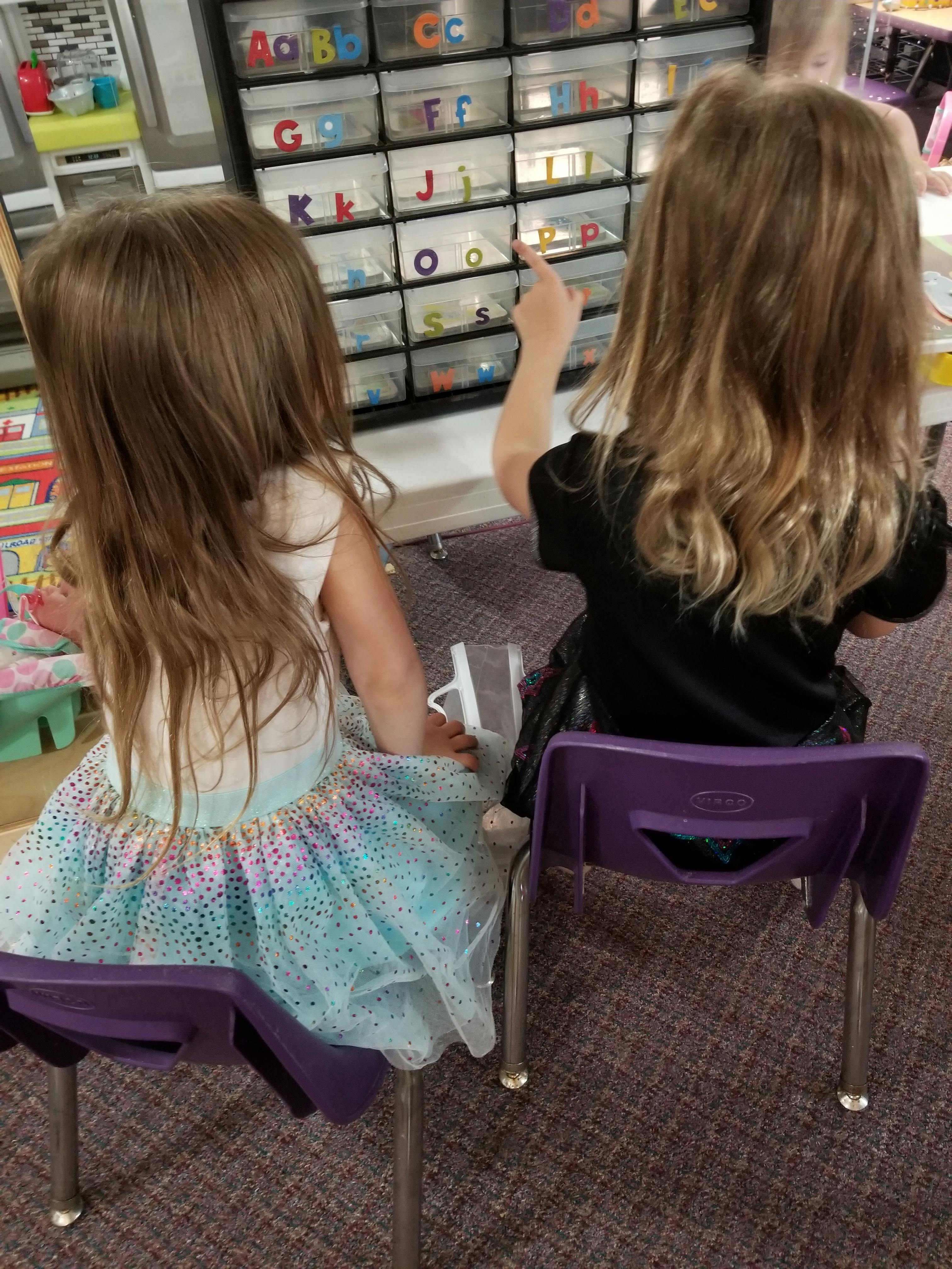 Two children sitting in chairs, one pointing at alphabet letters in a classroom