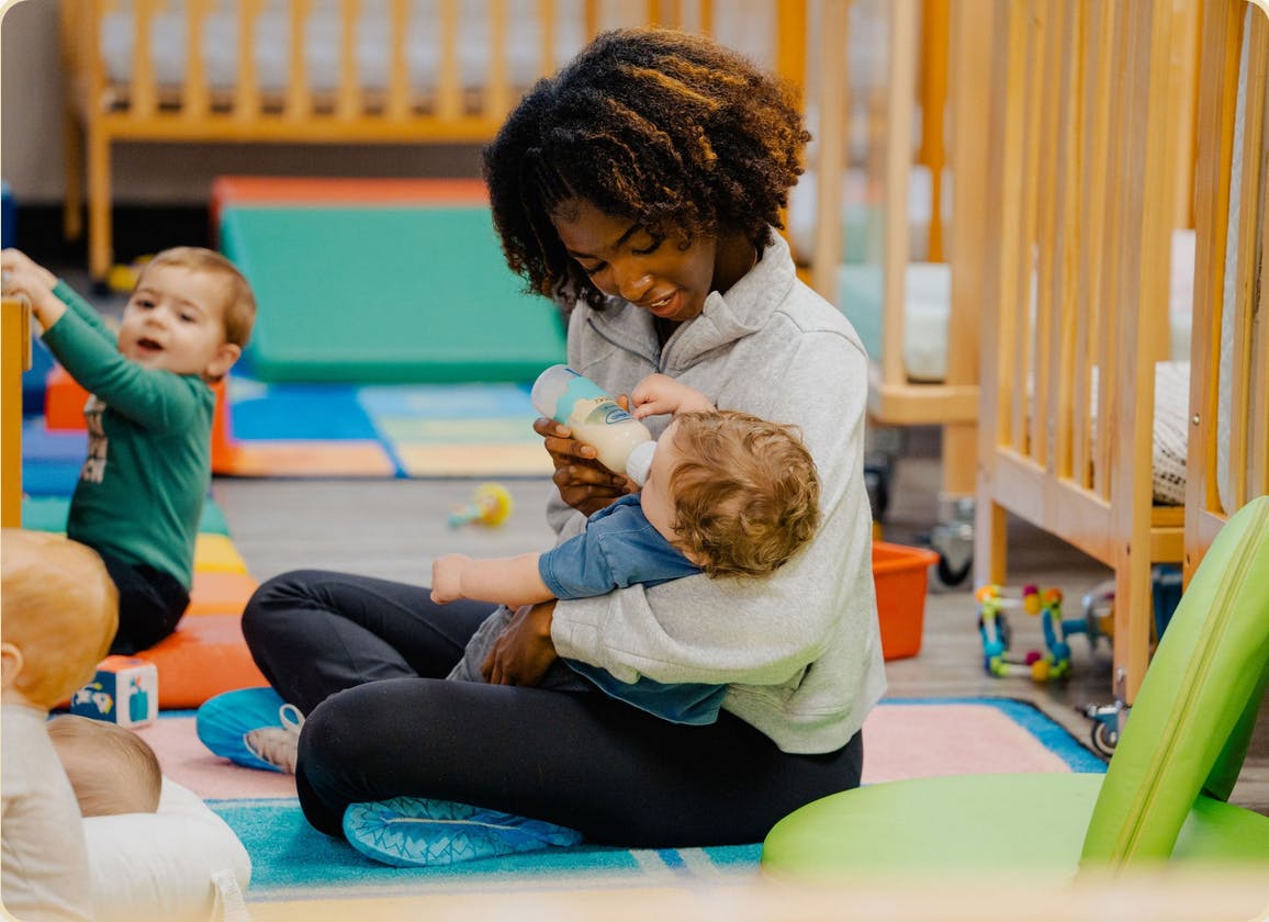 Person bottle-feeding a baby while sitting on the floor in a room with cribs