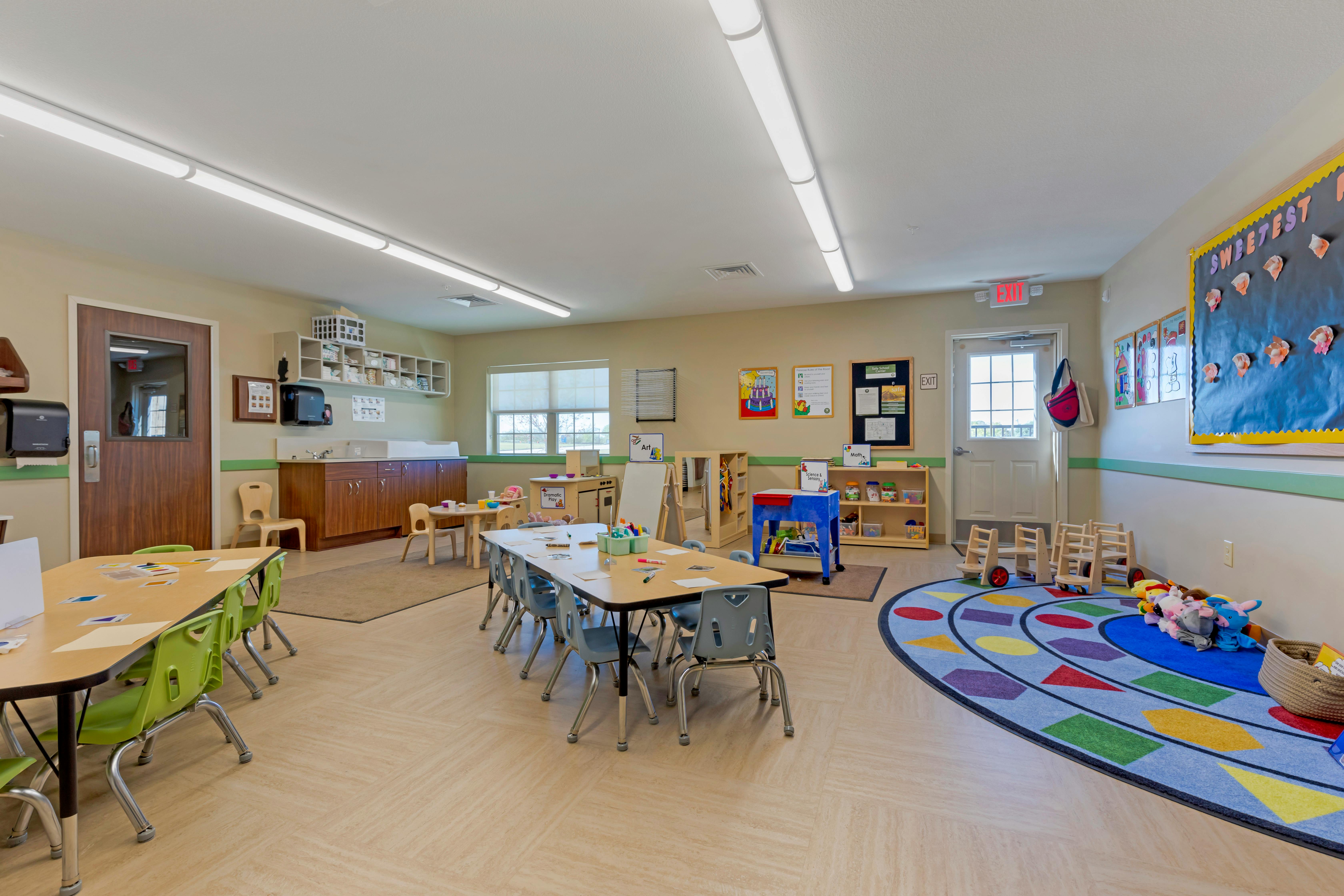 Classroom with multiple tables and chairs, a play area, and an exit door