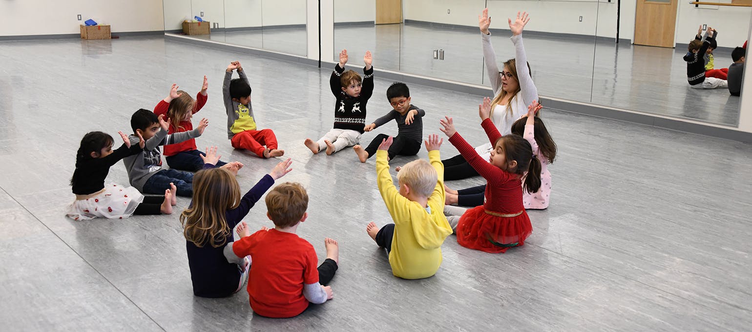 Children and an adult sitting in a circle with arms raised in a dance studio