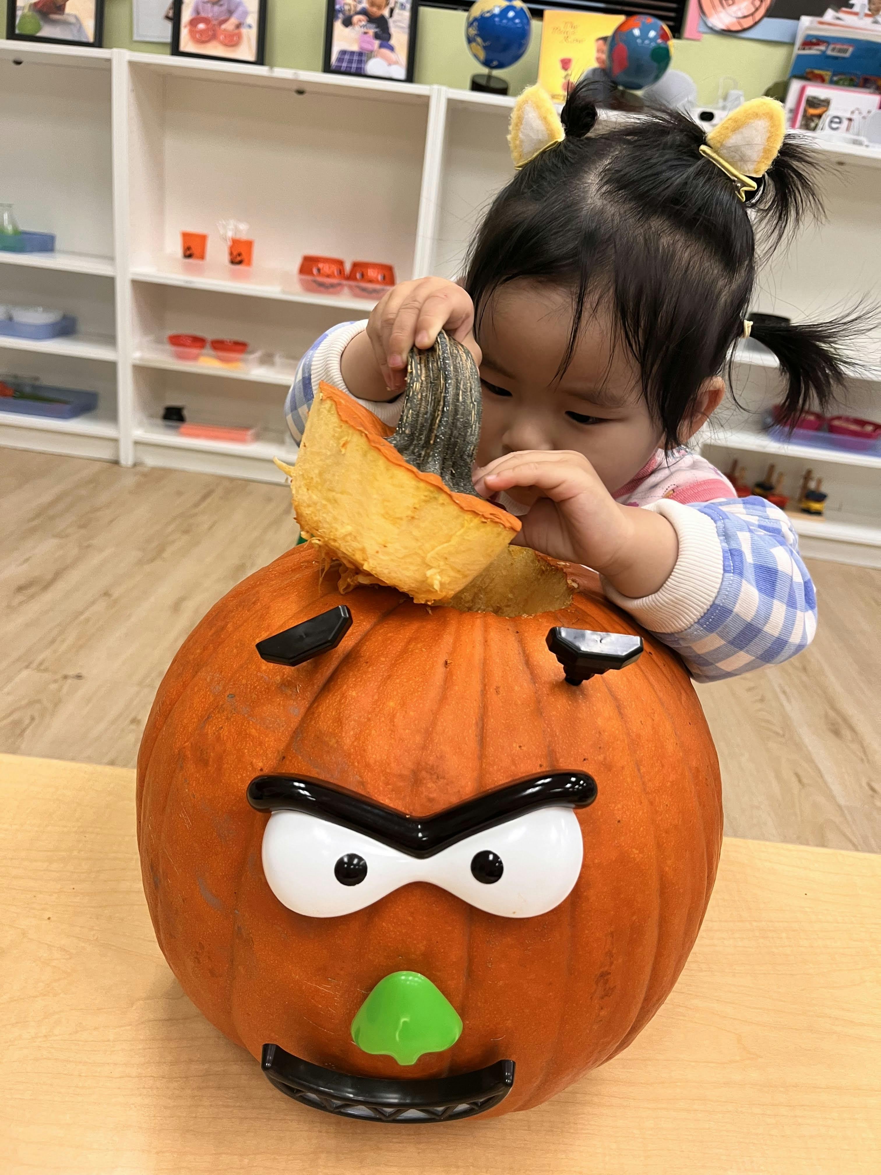 Child wearing cat ear hair clips, putting a top onto a pumpkin with plastic face parts