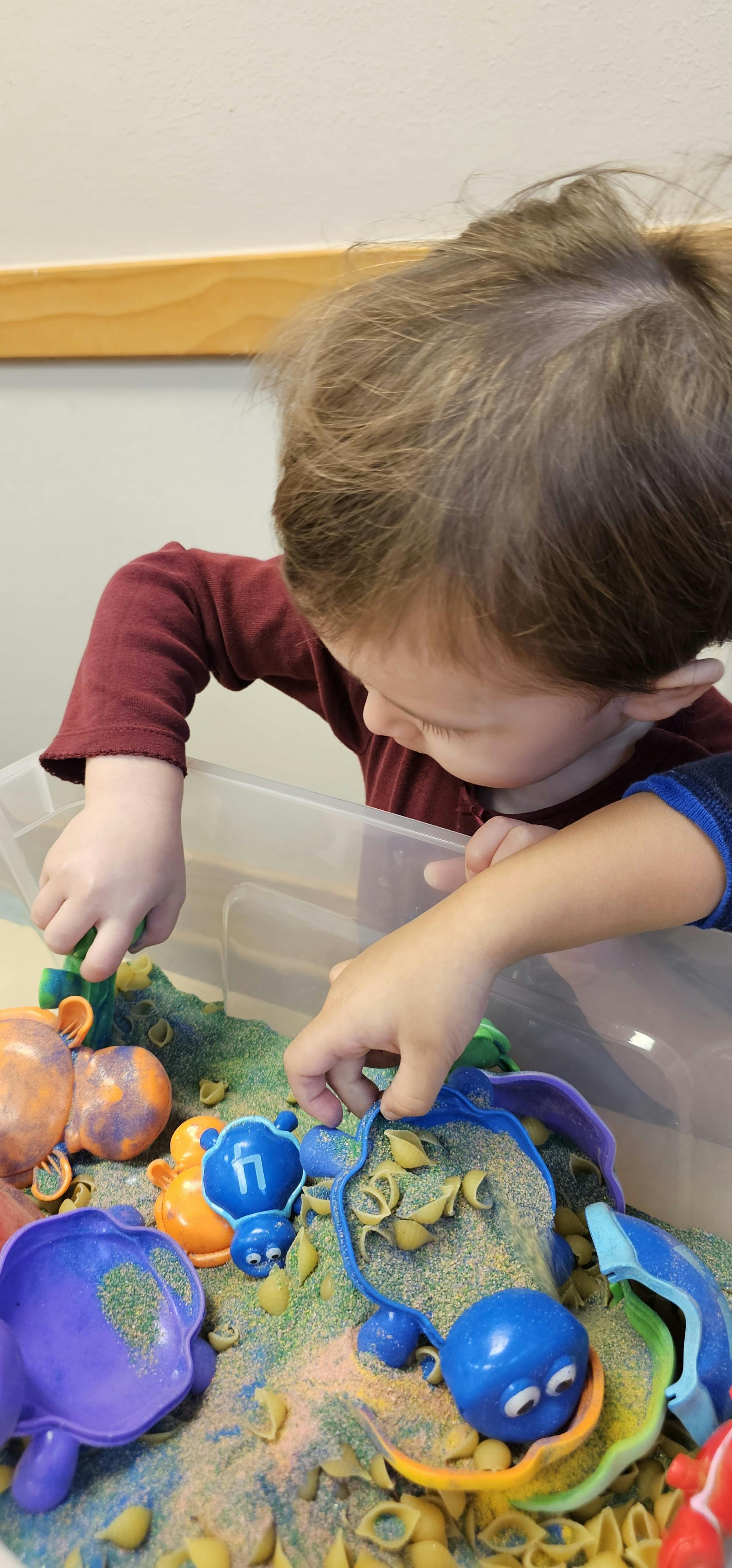Child playing with sand and pasta in a clear bin