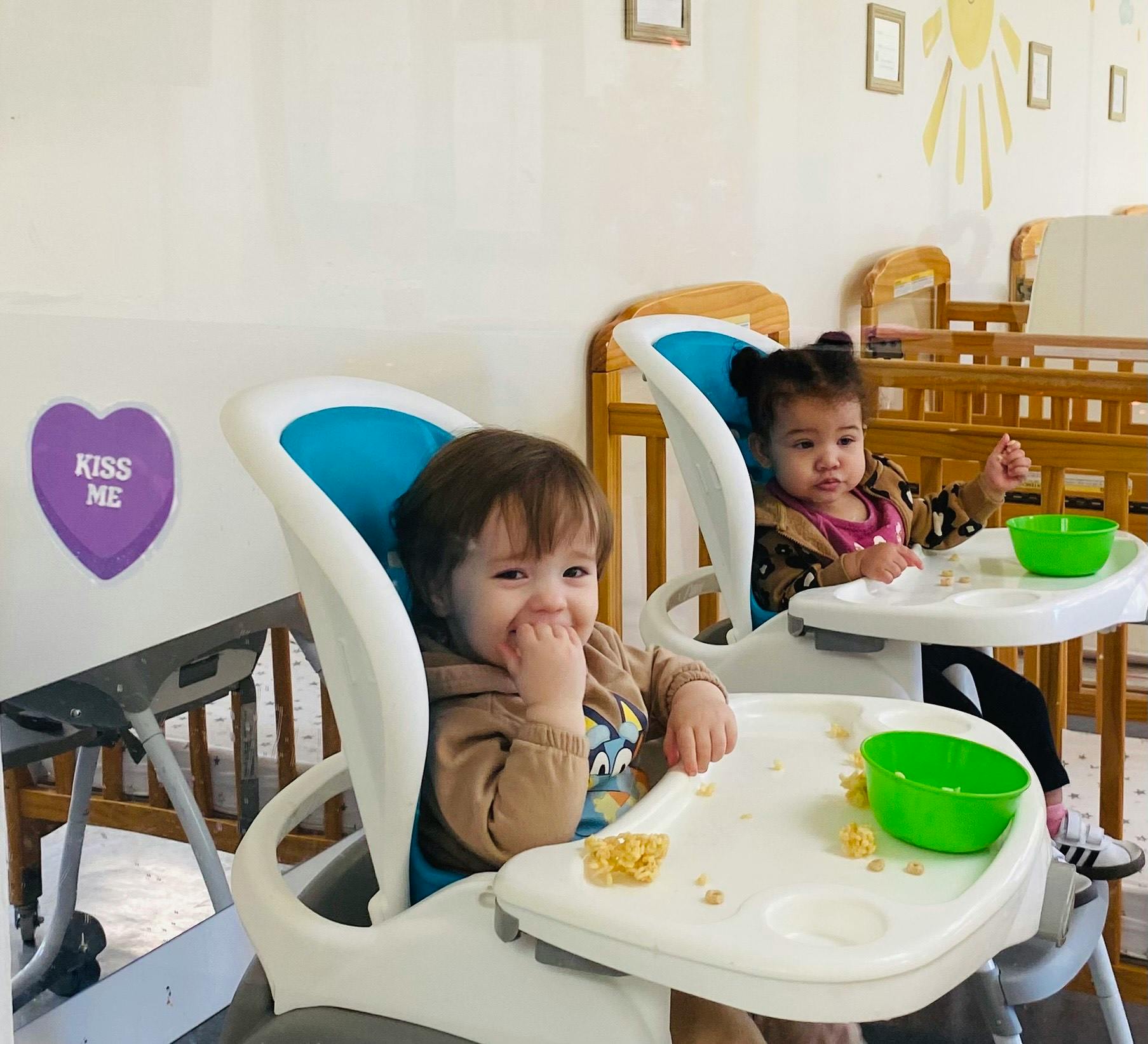 Two children in high chairs with snacks, one smiling and one looking ahead