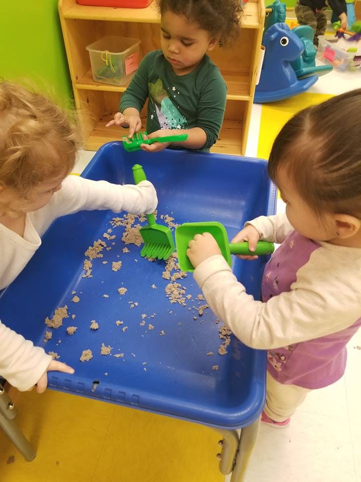 Three children playing with sand and green shovels in a blue bin