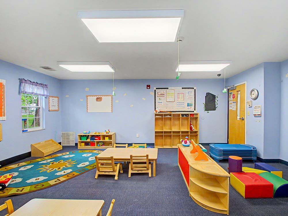 Classroom with small tables and chairs, a play rug, and shelves with toys