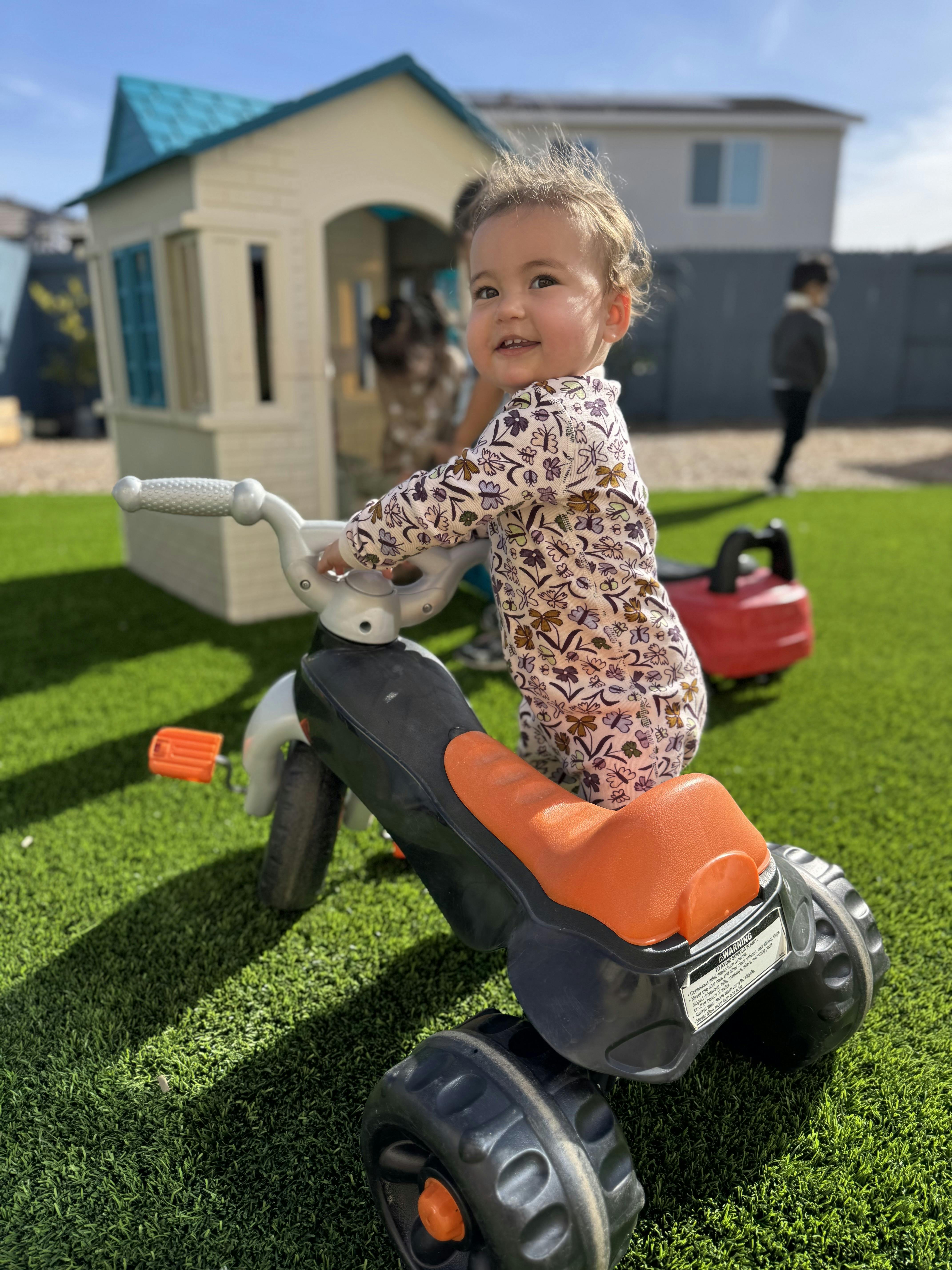 Child standing by a tricycle on artificial grass, smiling