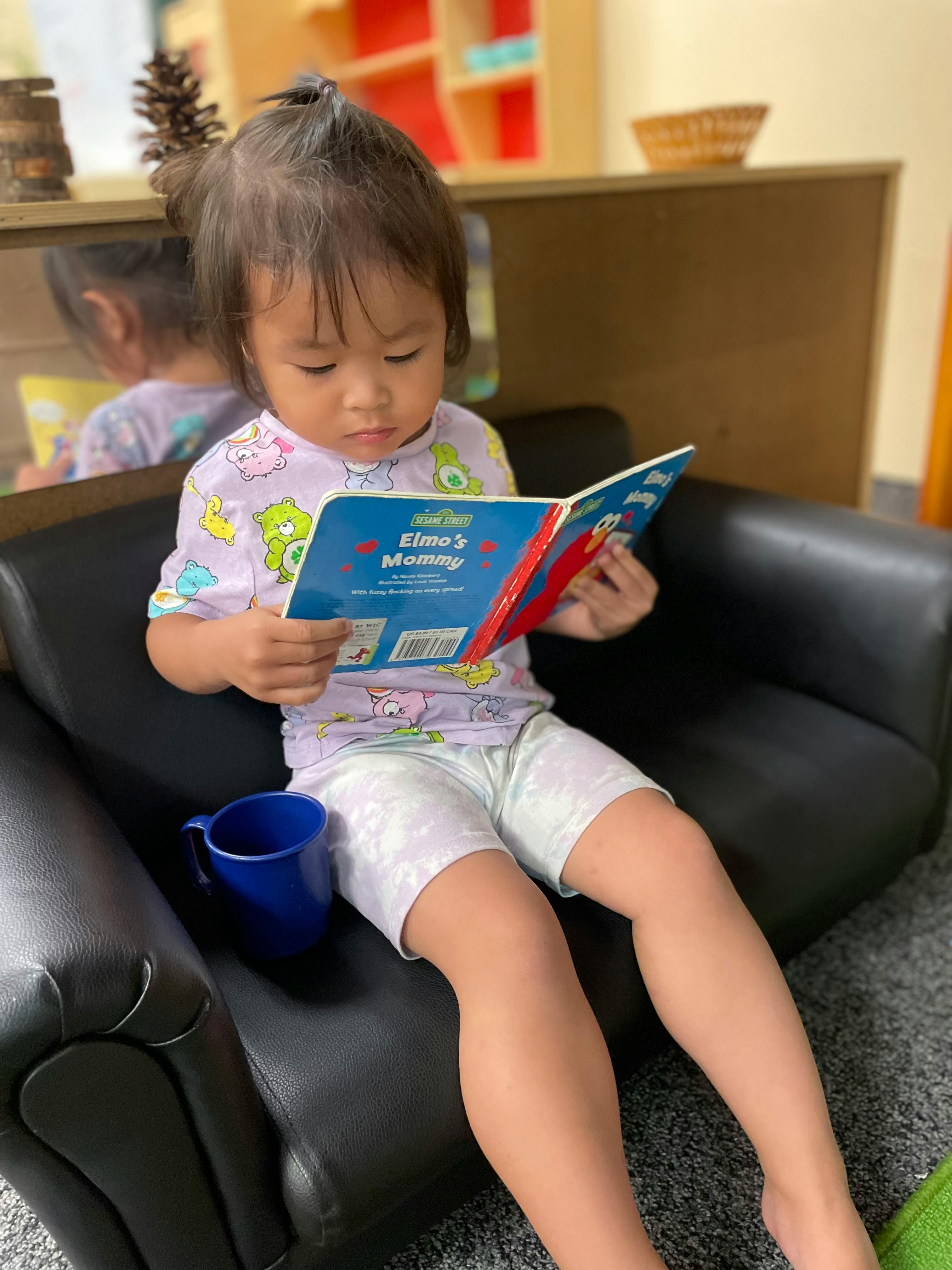 Child sitting on a chair reading a book titled "Elmo's Mommy"