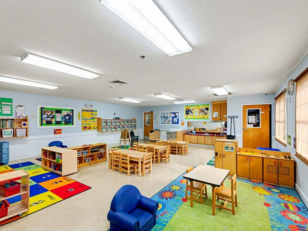 Empty classroom with small tables and chairs, colorful rugs, and learning materials