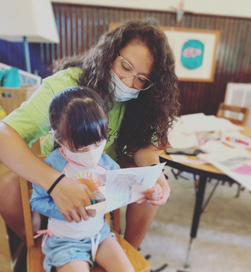 Person in mask helping child in mask read a paper