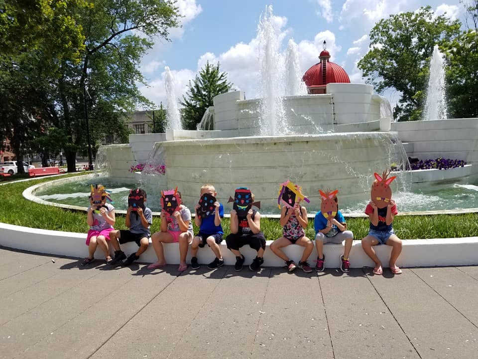 Seven children sitting in front of a fountain, holding masks over their faces