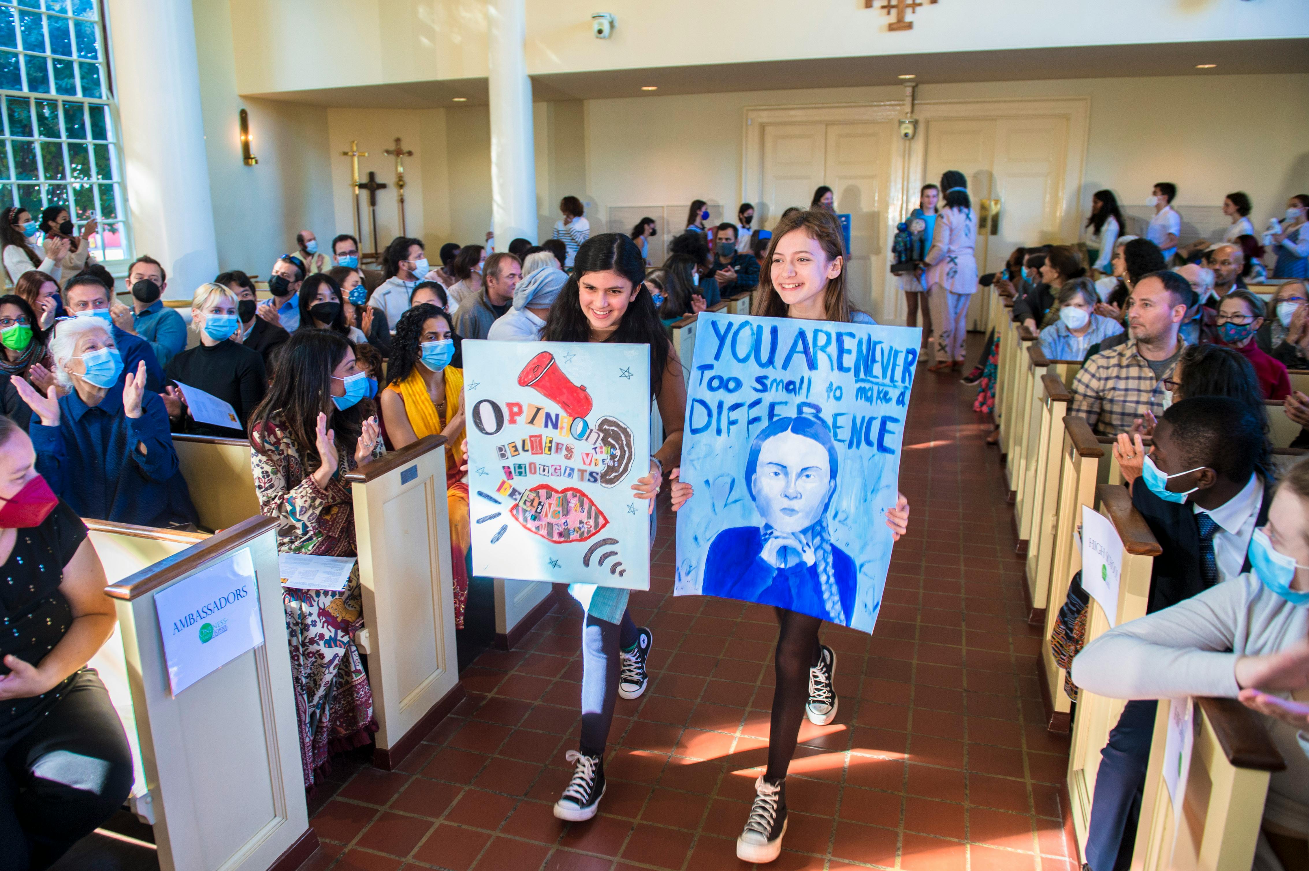 Two teens holding signs walking down an aisle past seated people