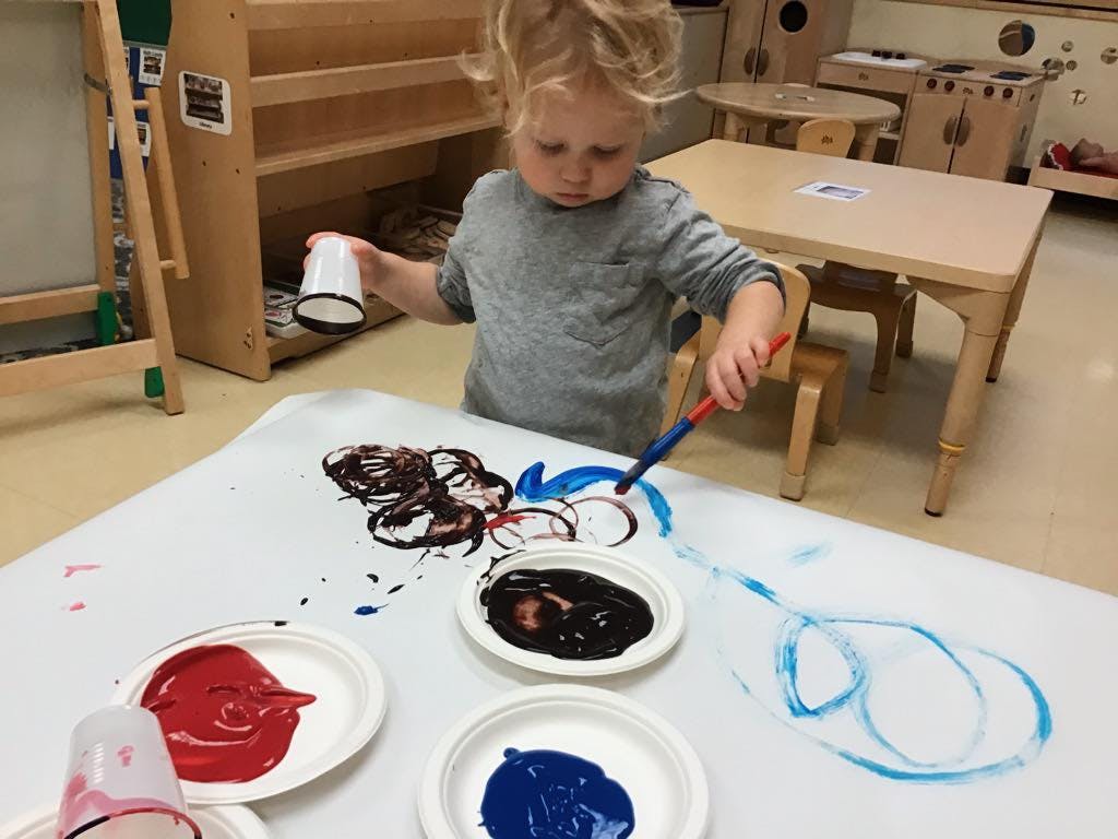 Child painting on paper with a brush and holding a cup