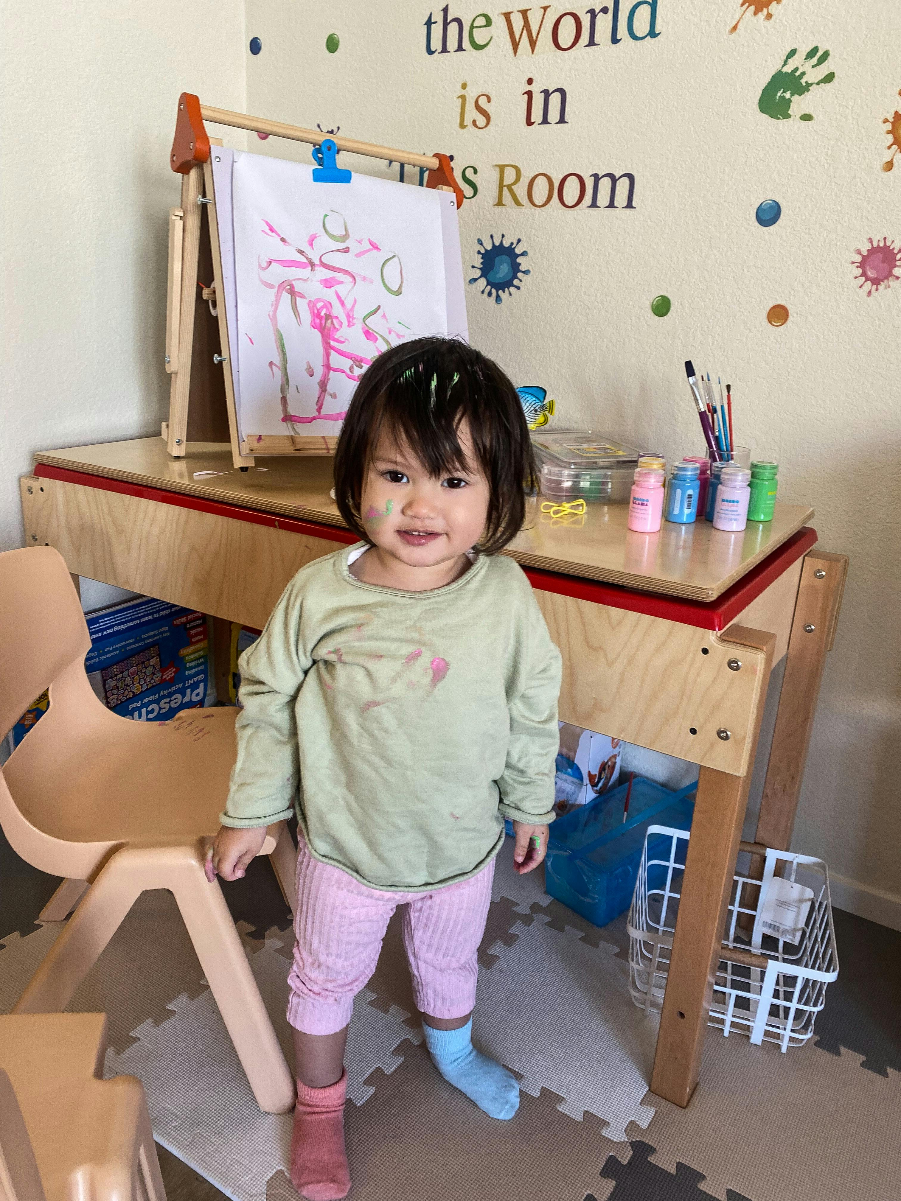Child covered in paint standing in front of an easel with a painting