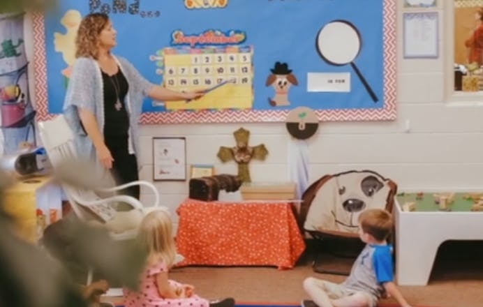 Person pointing at a bulletin board with children sitting on the floor