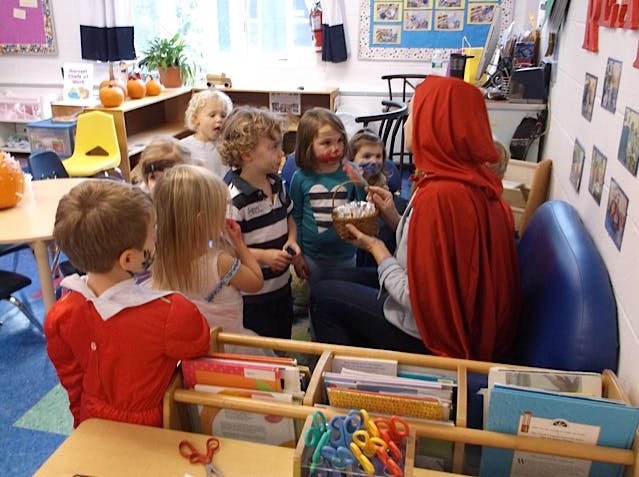 Person in red cape holding a cake for children in a classroom
