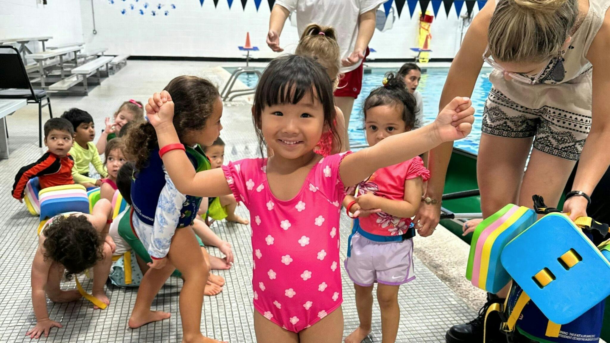 Child in pink swimsuit smiling with other children and adults in a pool area