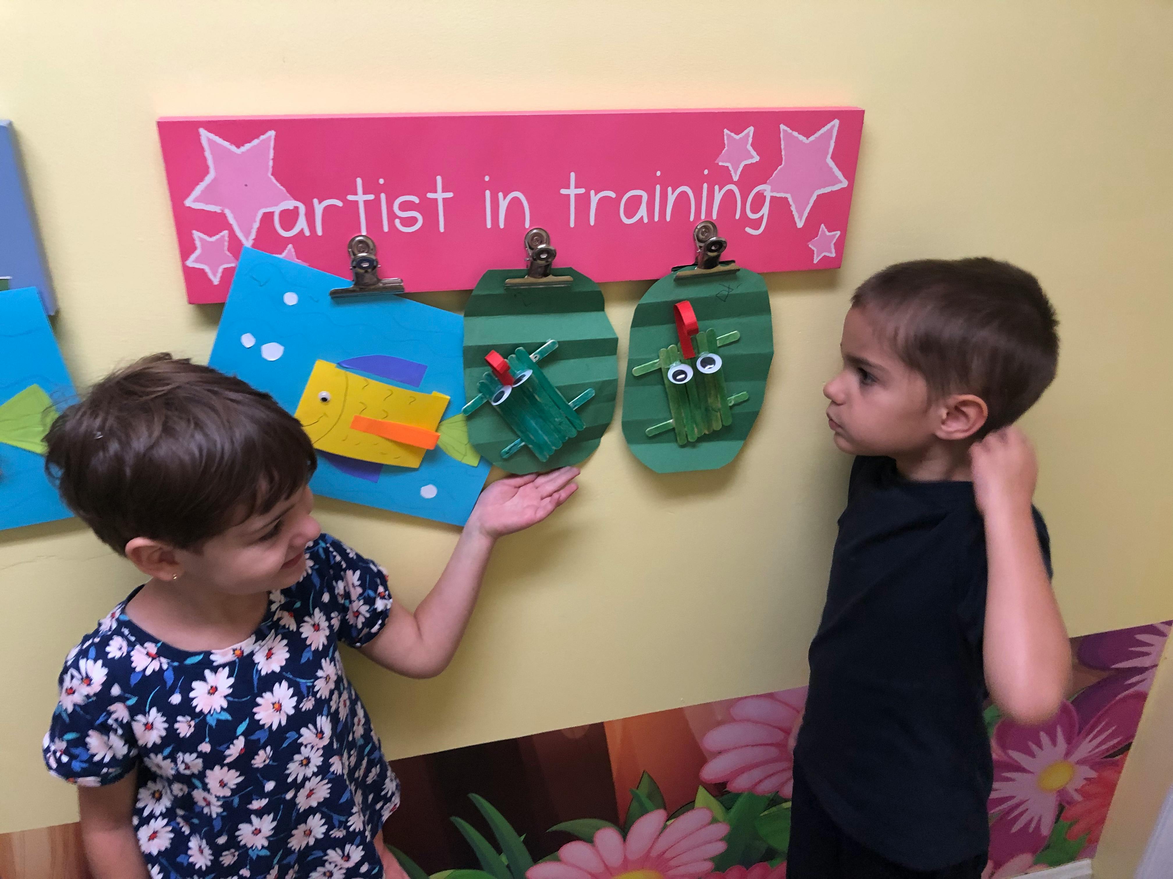 Two children looking at crafts on a wall with a sign that says "artist in training"