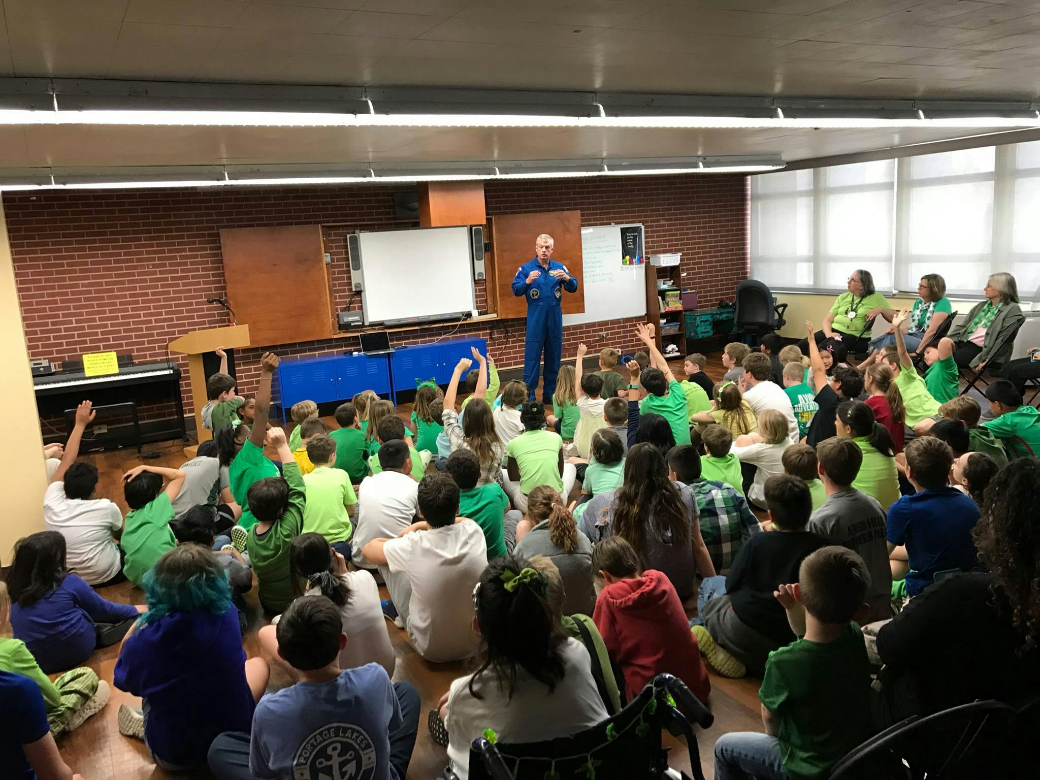 Person in a jumpsuit speaking to a group of children sitting on the floor