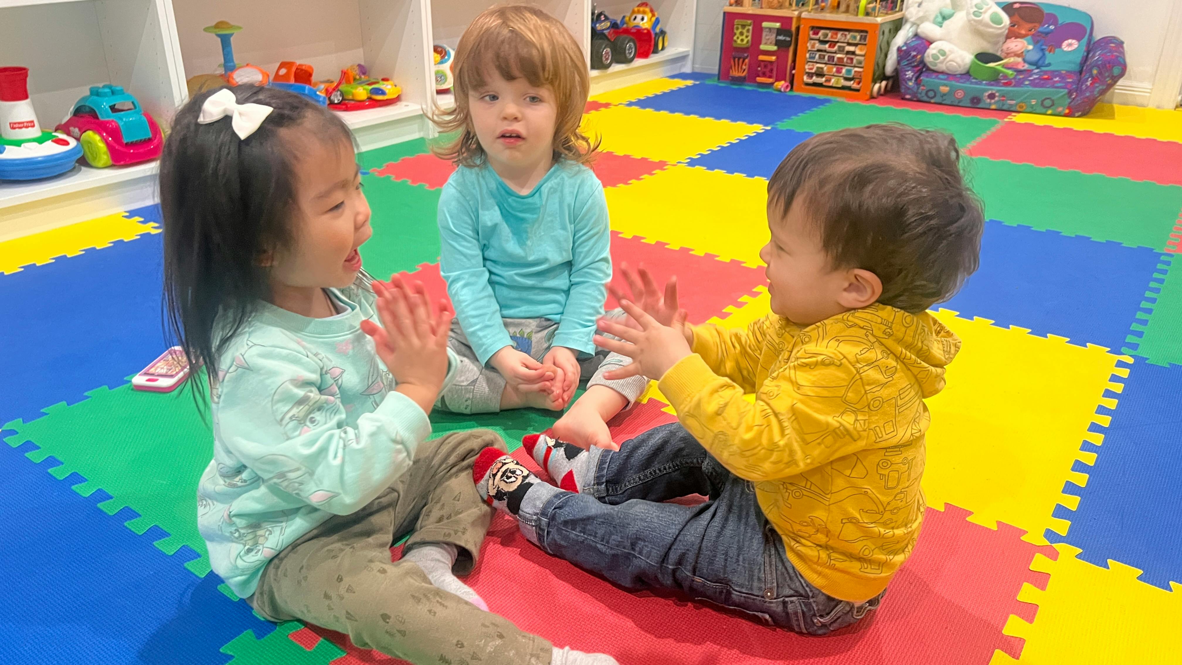 Three children sitting on a colorful mat, two clapping hands
