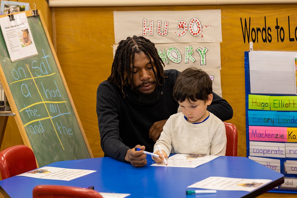 Person with dreadlocks helping a child write at a blue table in a classroom
