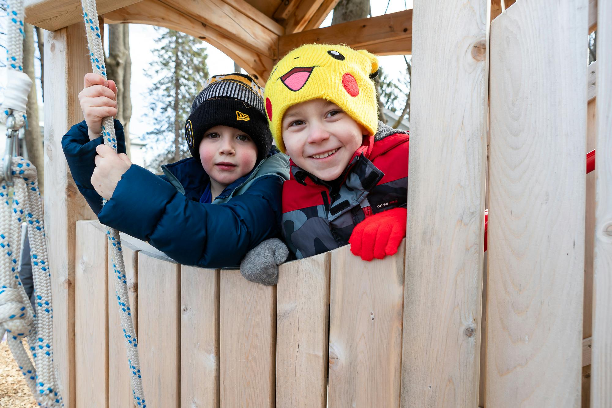 Two children in winter hats looking out from a wooden structure