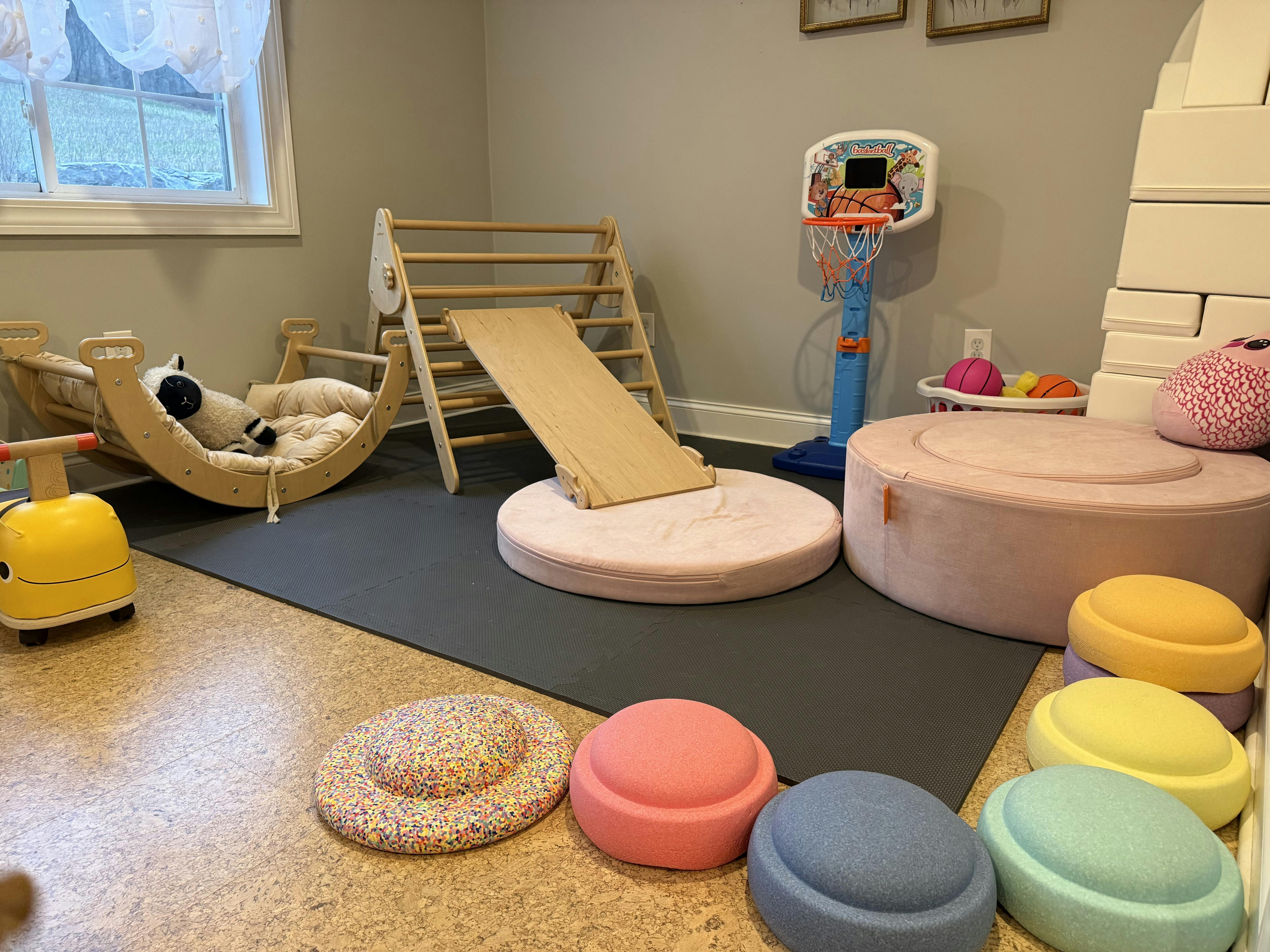 indoor playroom with wooden Pikler triangle, climbing arch, basketball hoop, and soft cushions
