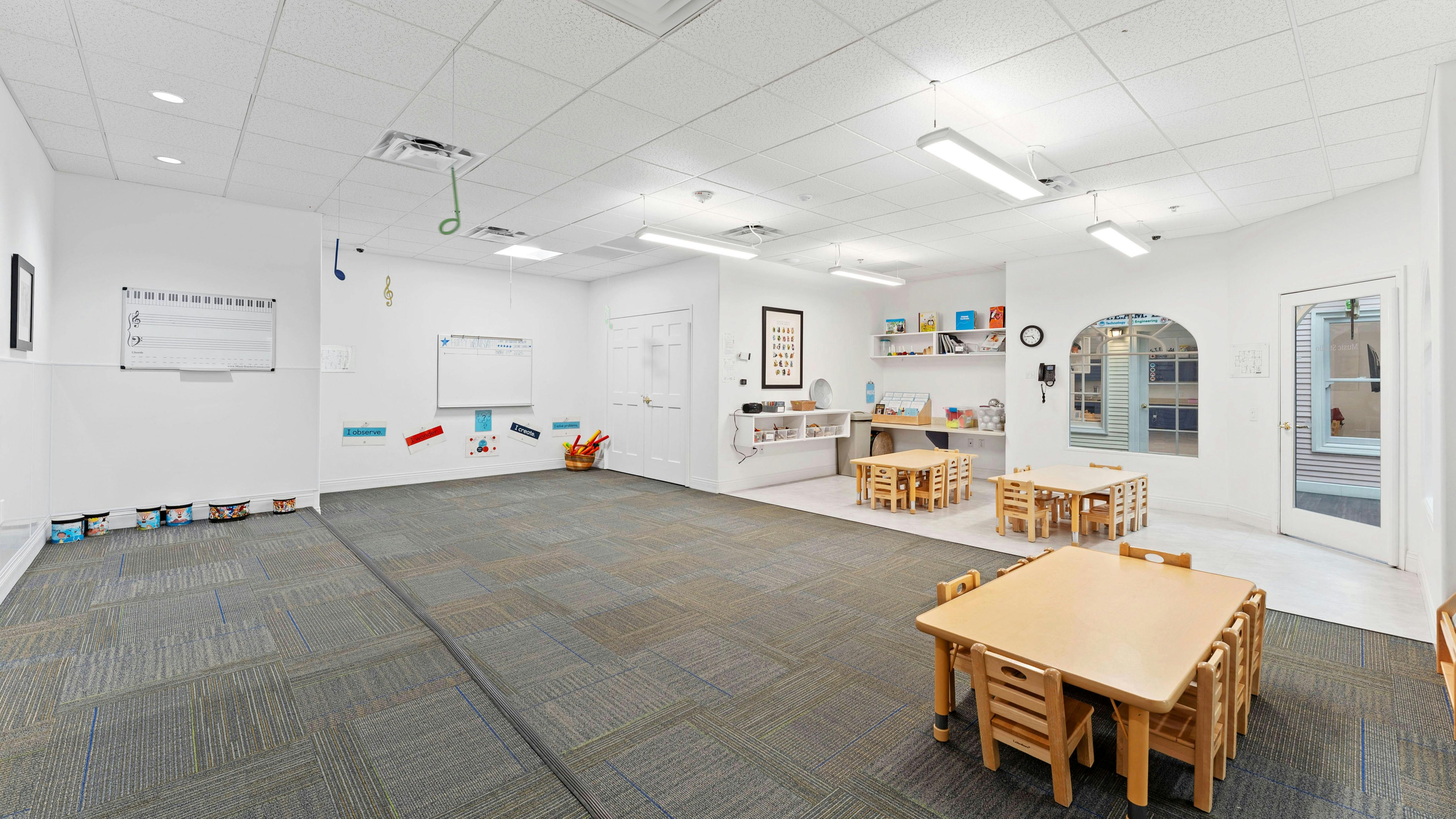 Classroom with small wooden tables and chairs, carpeted floor, and whiteboard with music notes