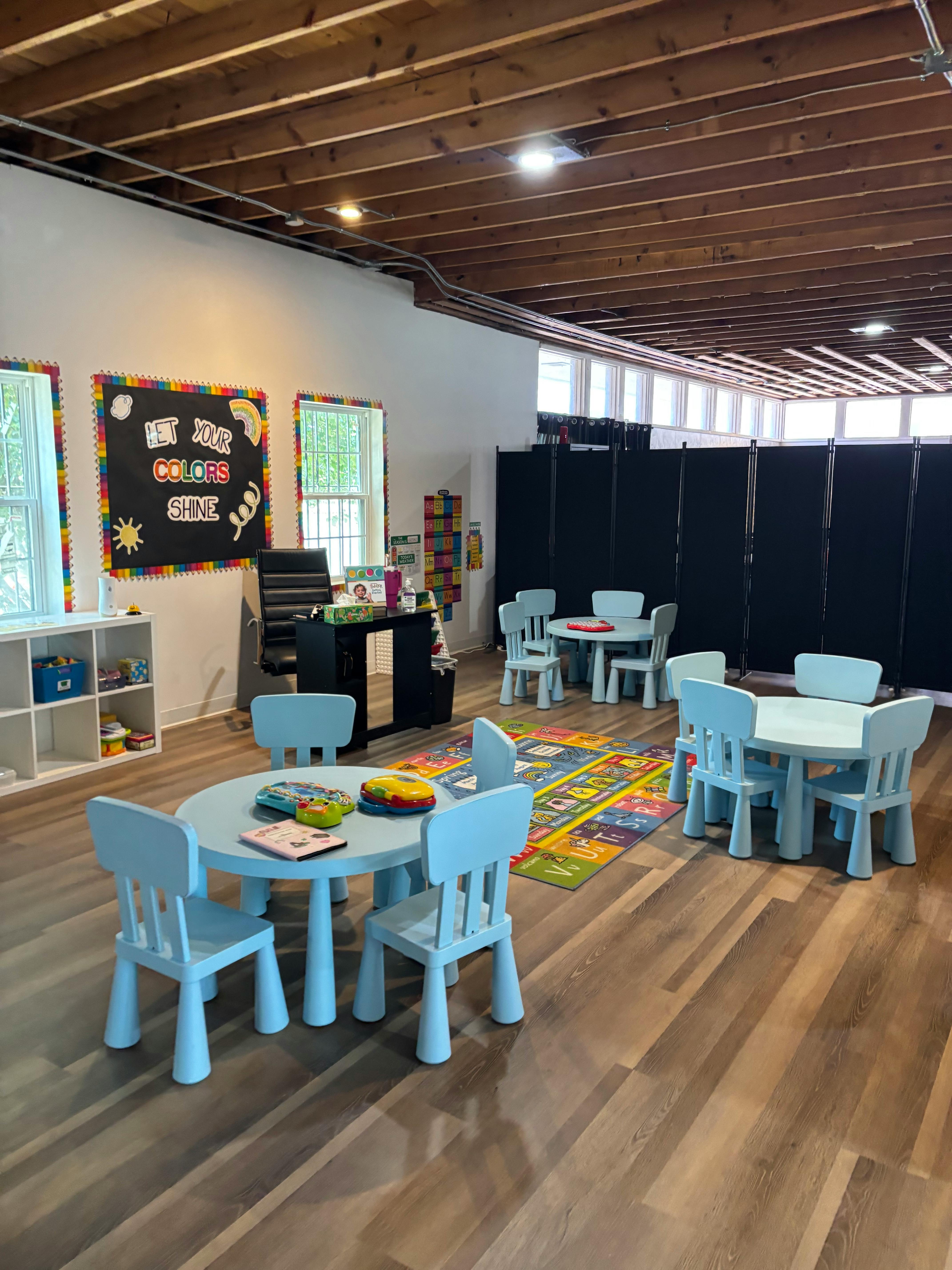 Classroom with small tables and chairs, colorful rug, and a blackboard with text