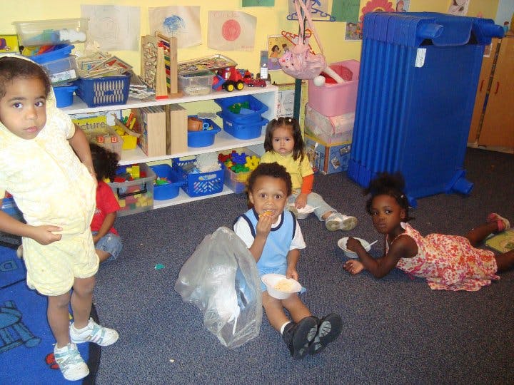 Four children in a classroom, one standing, one sitting, two on the floor