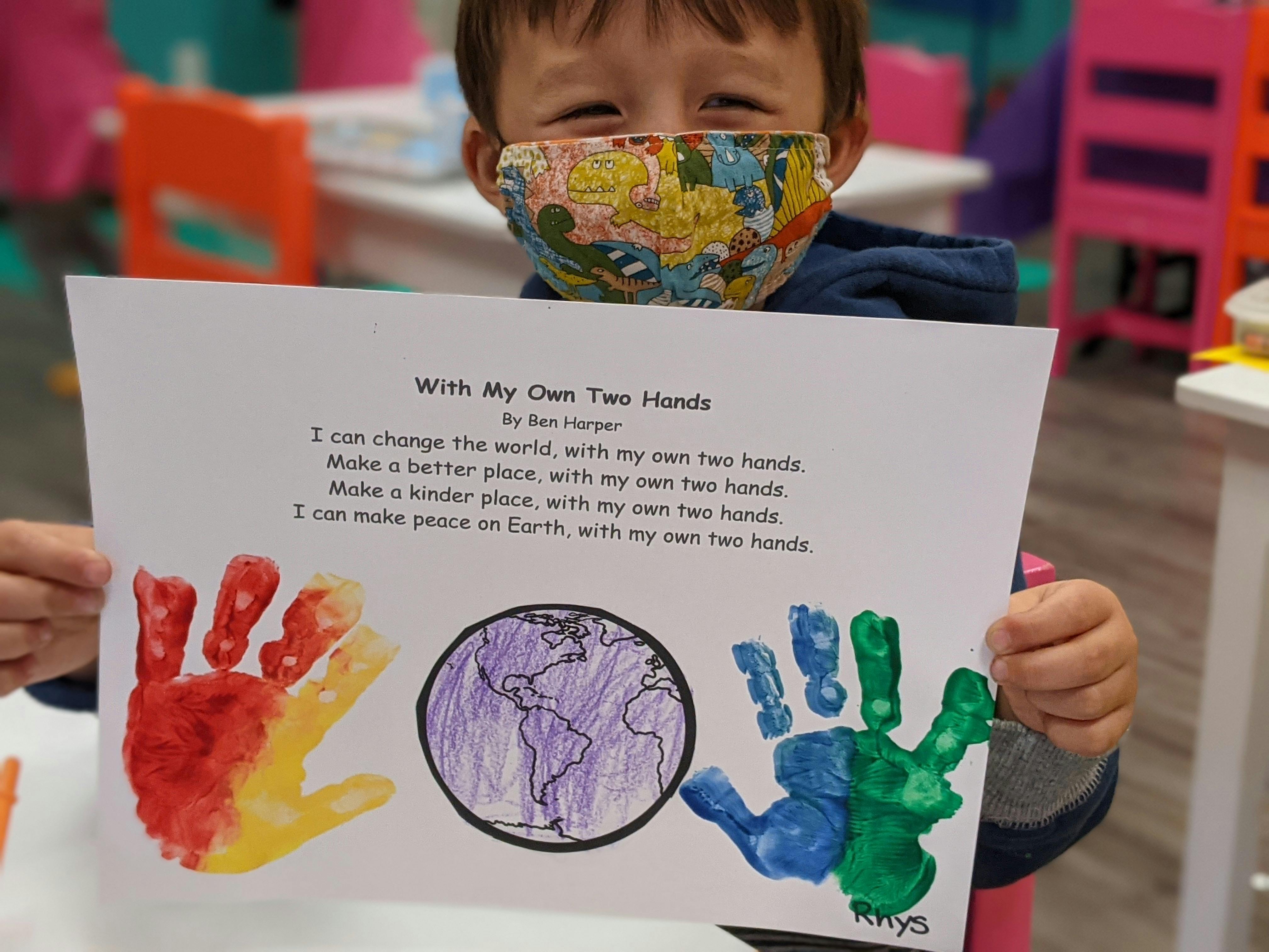 Child in a mask holding up a paper with handprints and a drawing of Earth