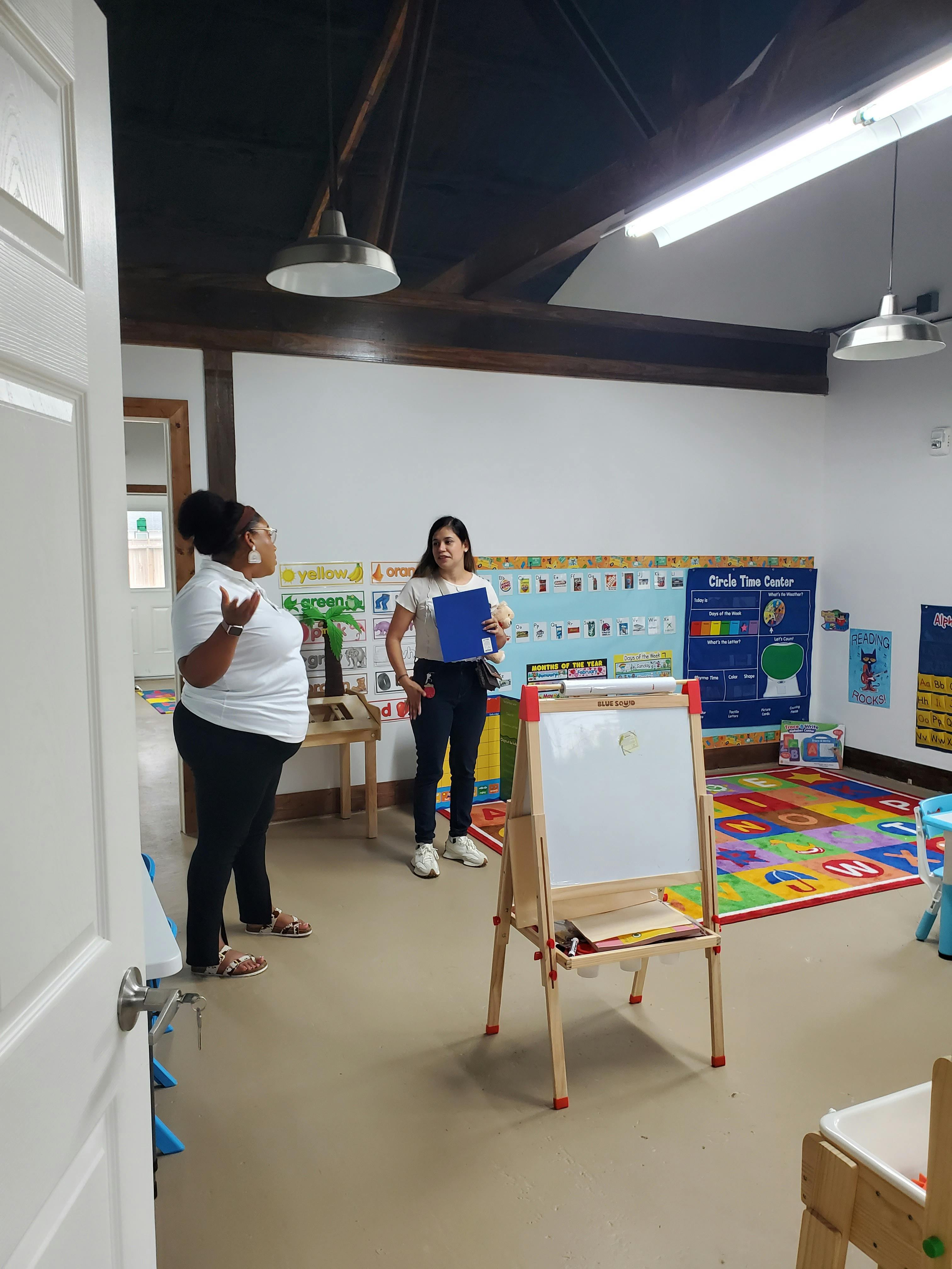 Two people standing in a classroom with an easel and colorful rug