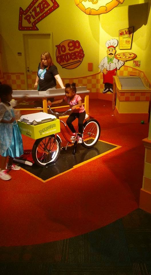 Child on a stationary bike with a delivery box, another child nearby